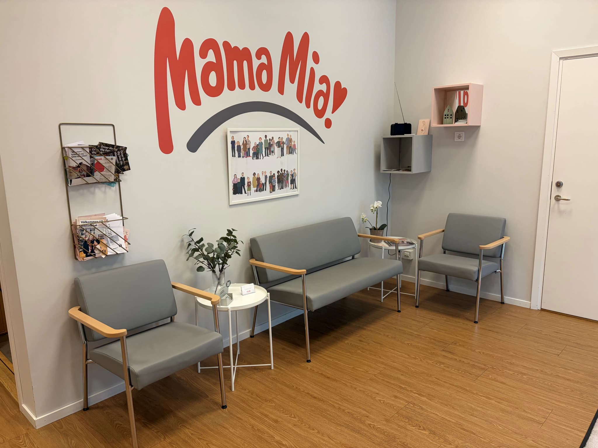 Väntrum med gråa fåtöljer, en soffa, ett runt vitt bord med en vas med gröna blad och en kaffekopp, samt väggdekorationer med texten "Mama Mia" och en bild på en familj.