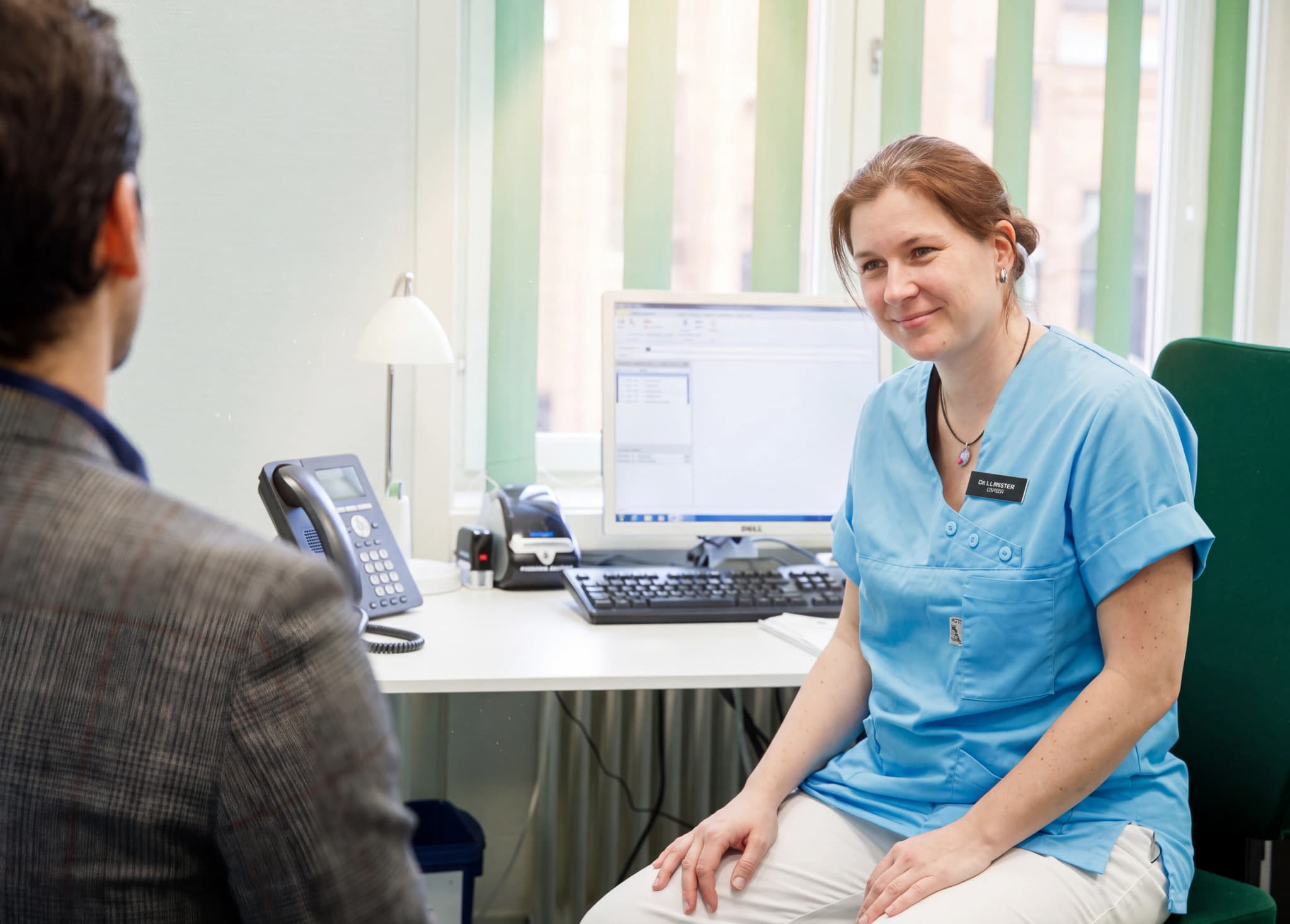 Sjuksköterska i ljusblå uniform ler mot en patient som sitter med ryggen mot kameran vid ett skrivbord med en telefon.
