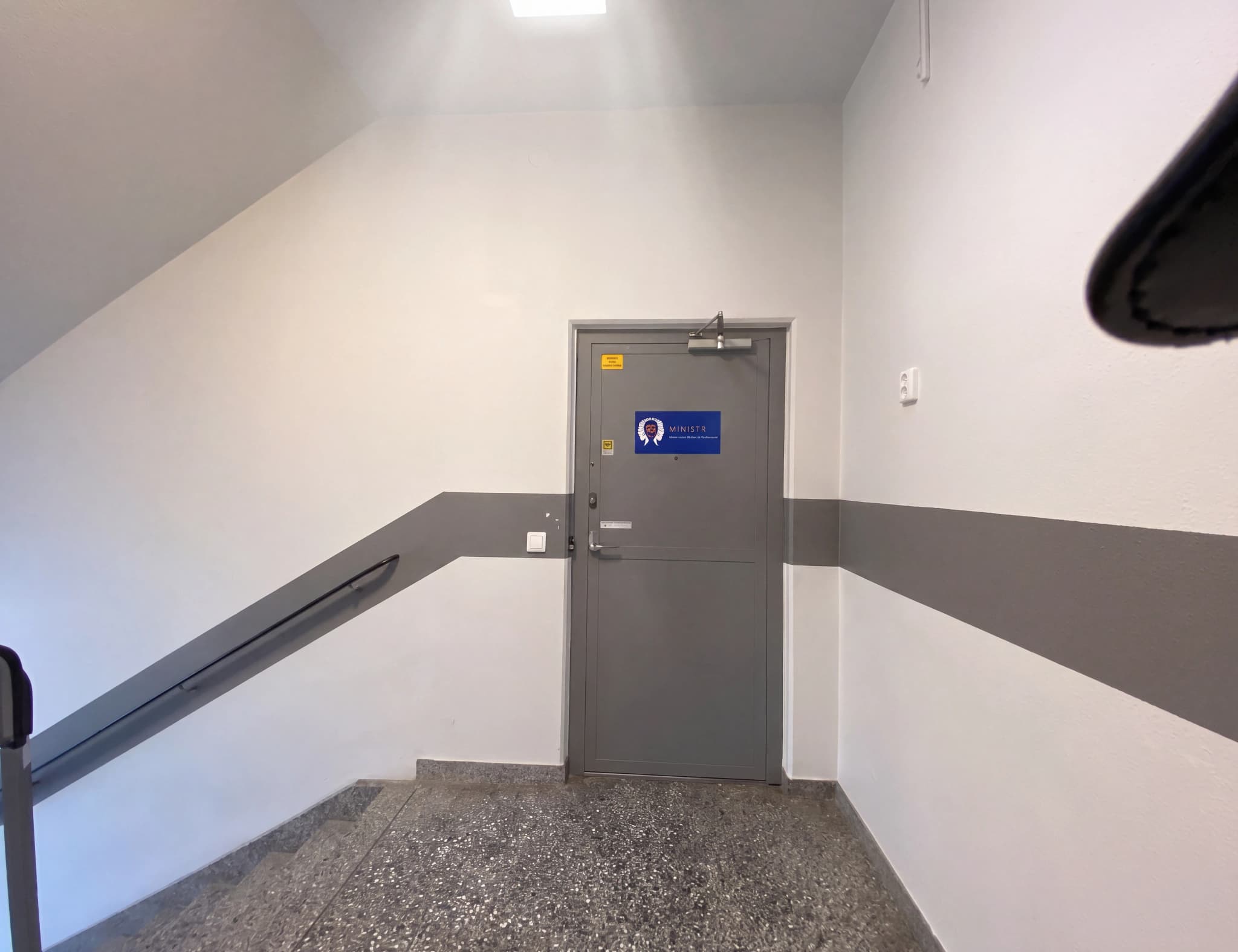 En interiörbild av en trappuppgång med en grå dörr och en horisontell grå rand längs väggen.