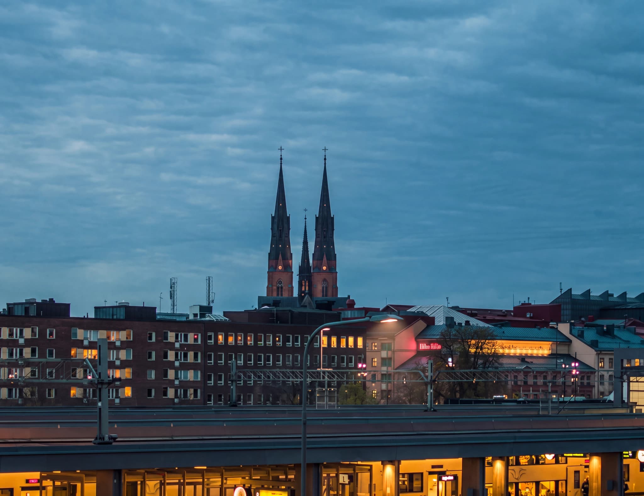 Kyrkspiror reser sig över stadens silhuett vid skymningen, med en molnig himmel och upplysta byggnader nedanför.