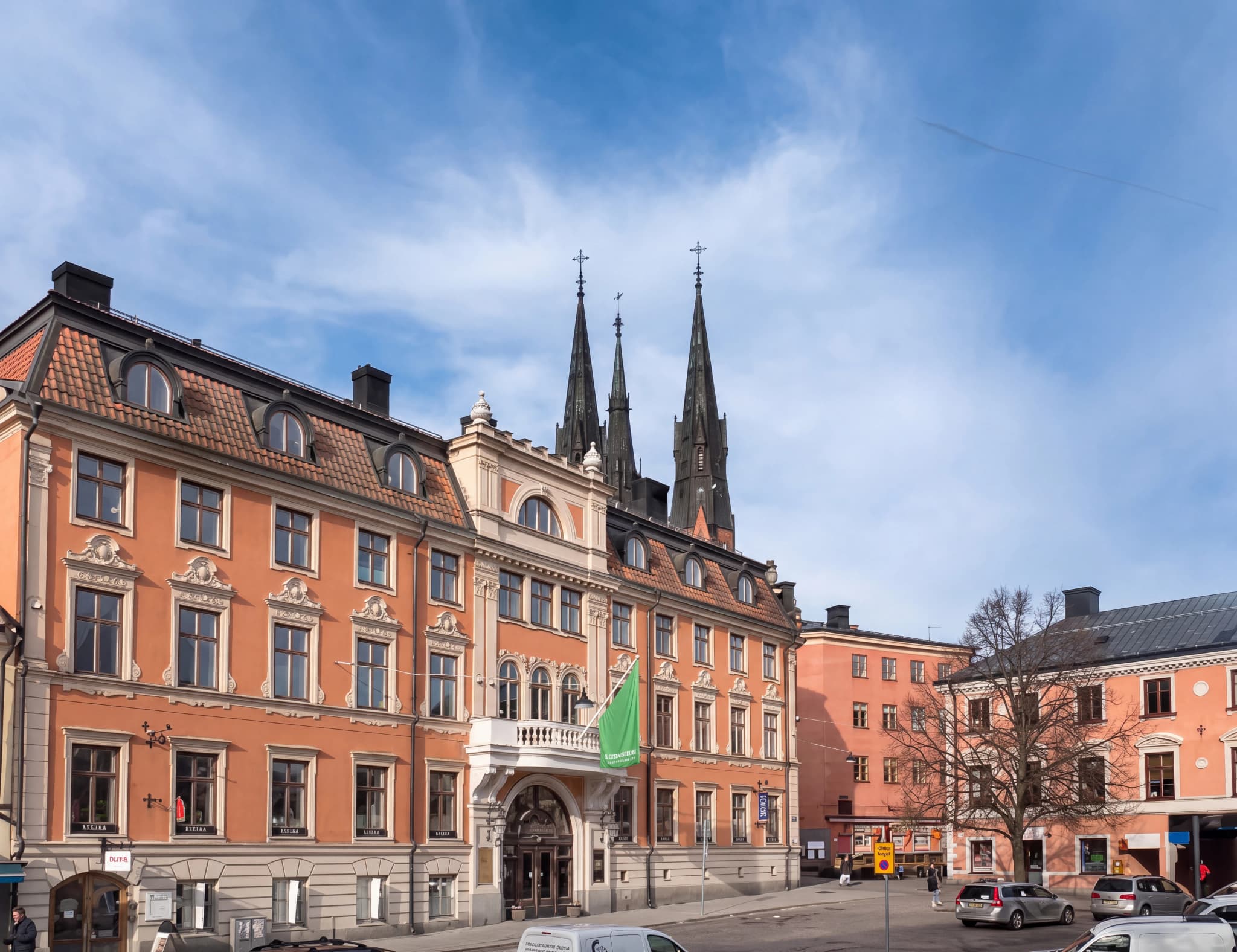 Fasaden av en orange byggnad med flera våningar och fönster, med två höga kyrkspiror som sticker upp bakom den på en blå, molnig himmel.