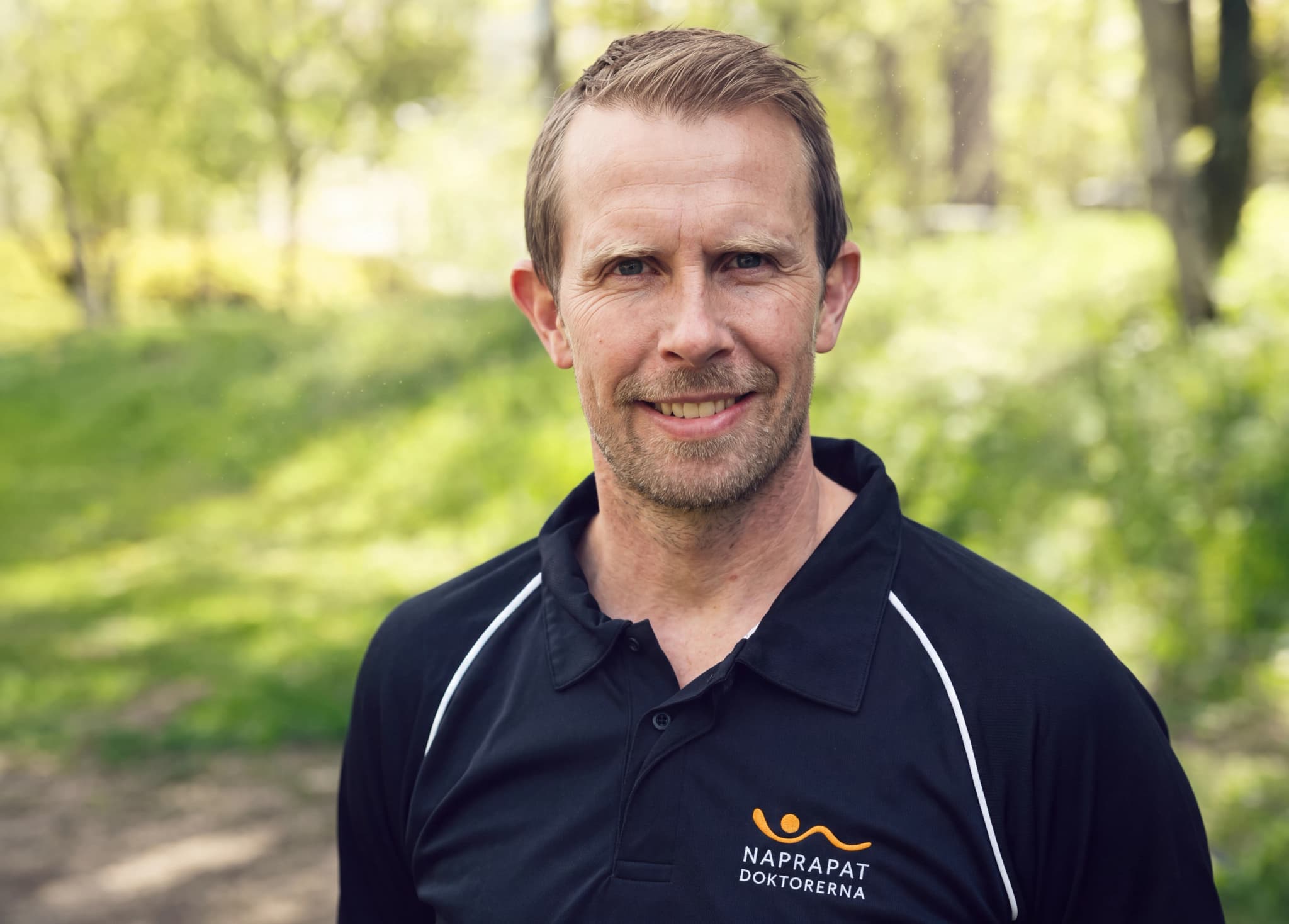 En man i svart pikétröja med Naprapat Doktorerna-logotypen ler mot kameran utomhus i en parkmiljö.