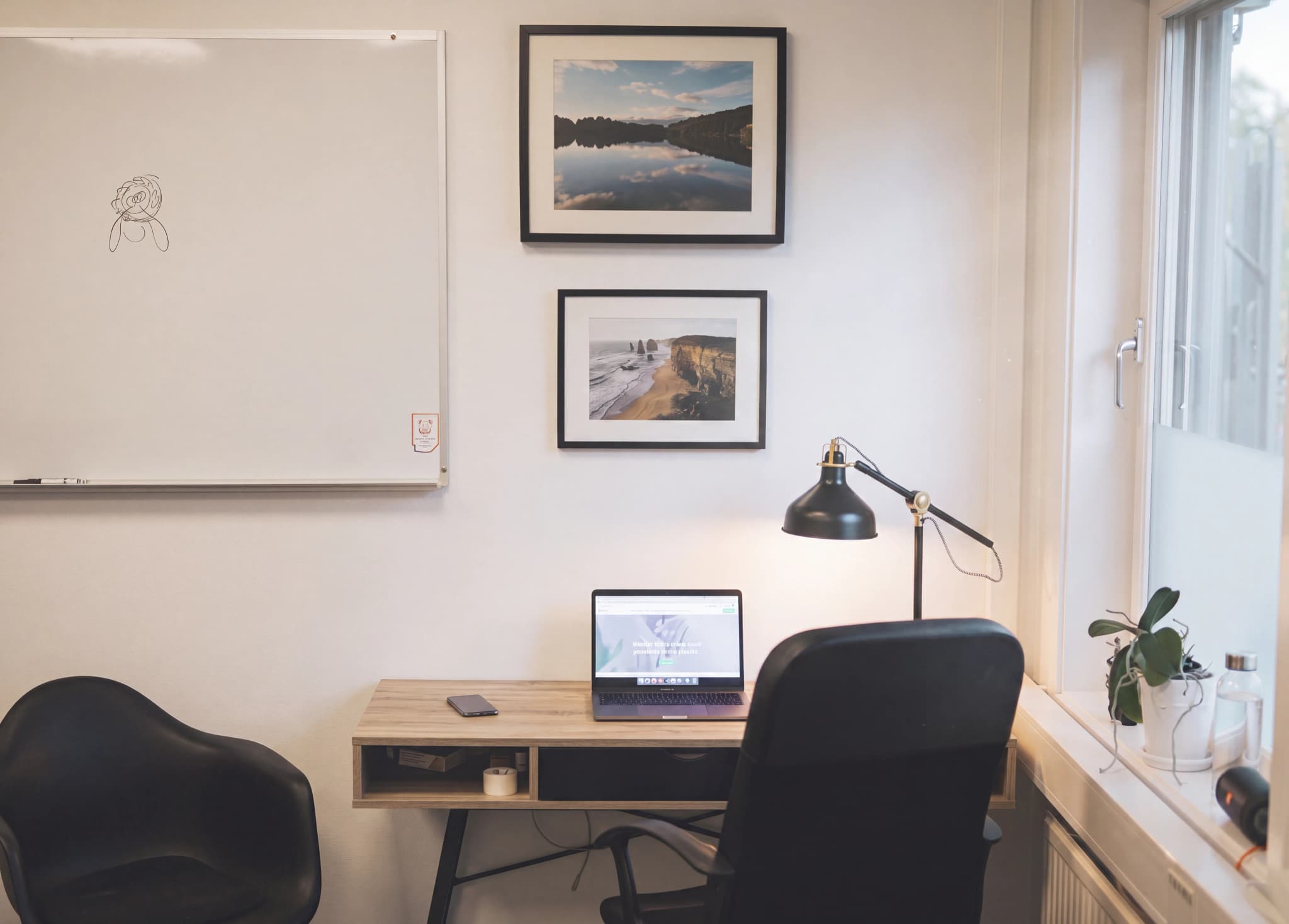 Ett modernt hemmakontor med ett ljust trä skrivbord, en laptop med en webbsida öppen, en svart kontorsstol och en vit whiteboardtavla med en enkel teckning.