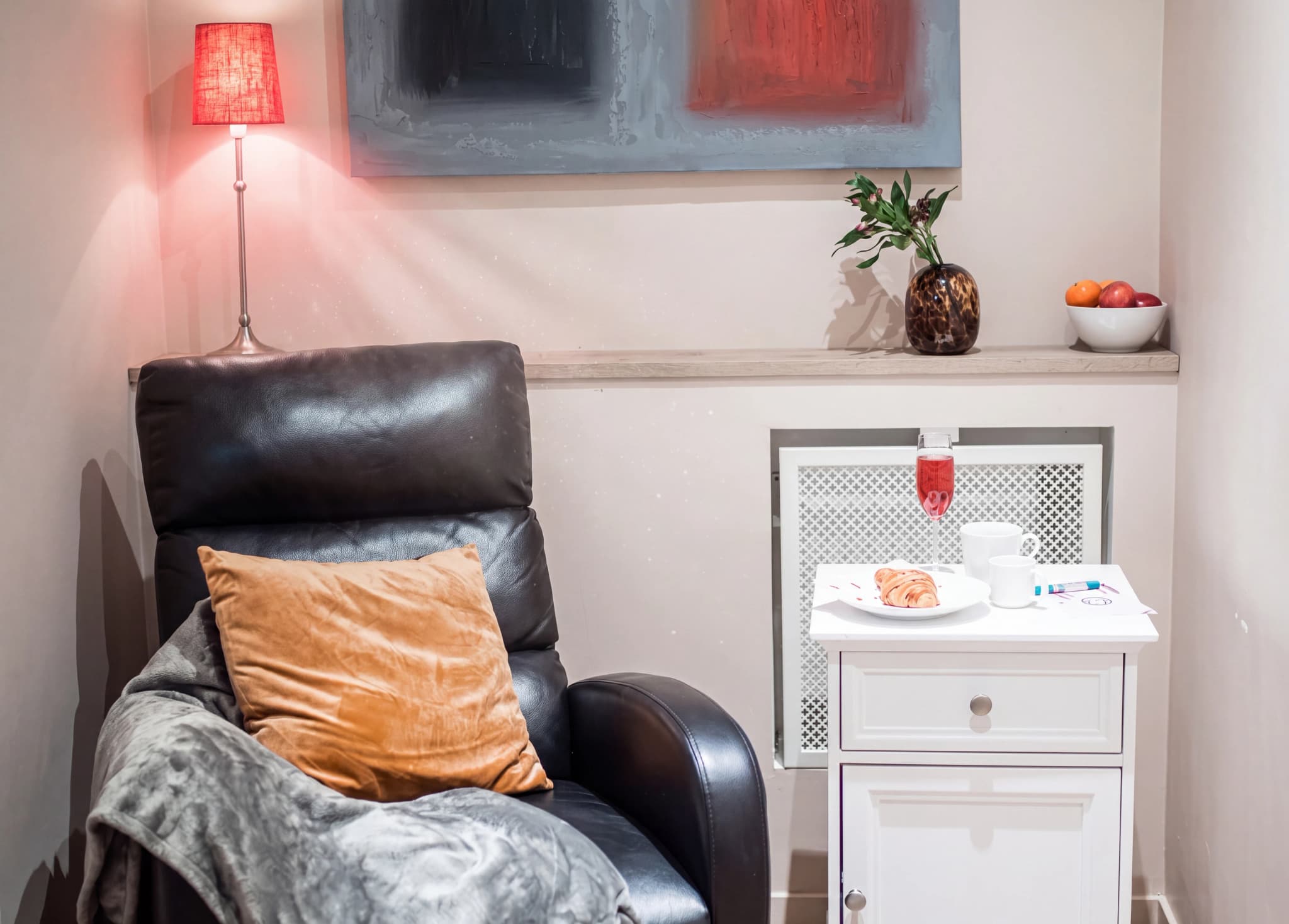 Bekväm svart reclinerfåtölj med en orange kudde och en filt, bredvid ett vitt nattduksbord med en bricka med en croissant, en kopp och ett glas röd dryck, samt en vas med blommor och en fruktskål på en hylla ovanför.