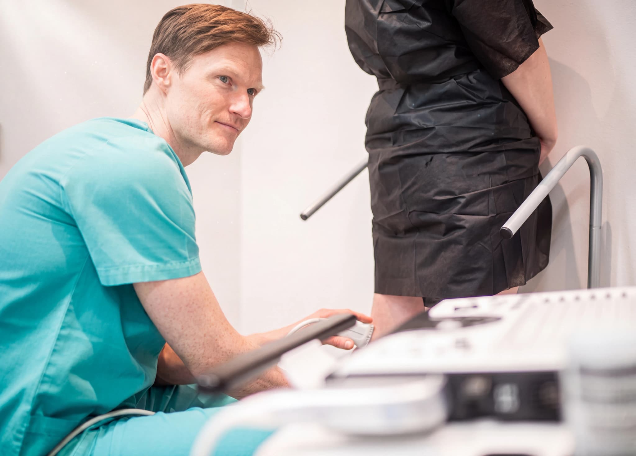 Läkare i grön skrubb undersöker en patients ben med en ultraljudsapparat i ett kliniskt rum.