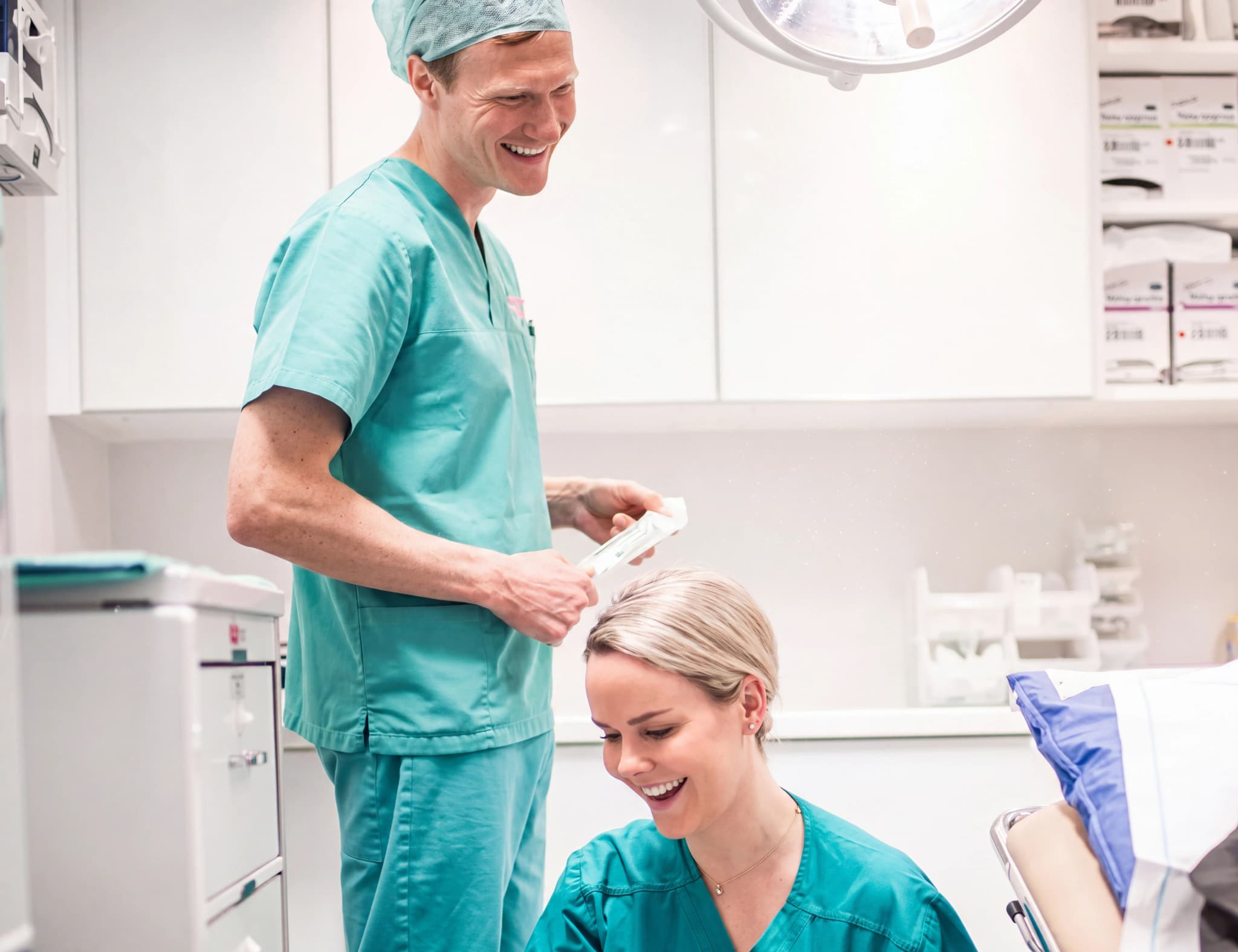 Två läkare i turkos sjukhusklädsel ler medan de tittar på och hanterar medicinska förnödenheter i ett kliniskt rum.
