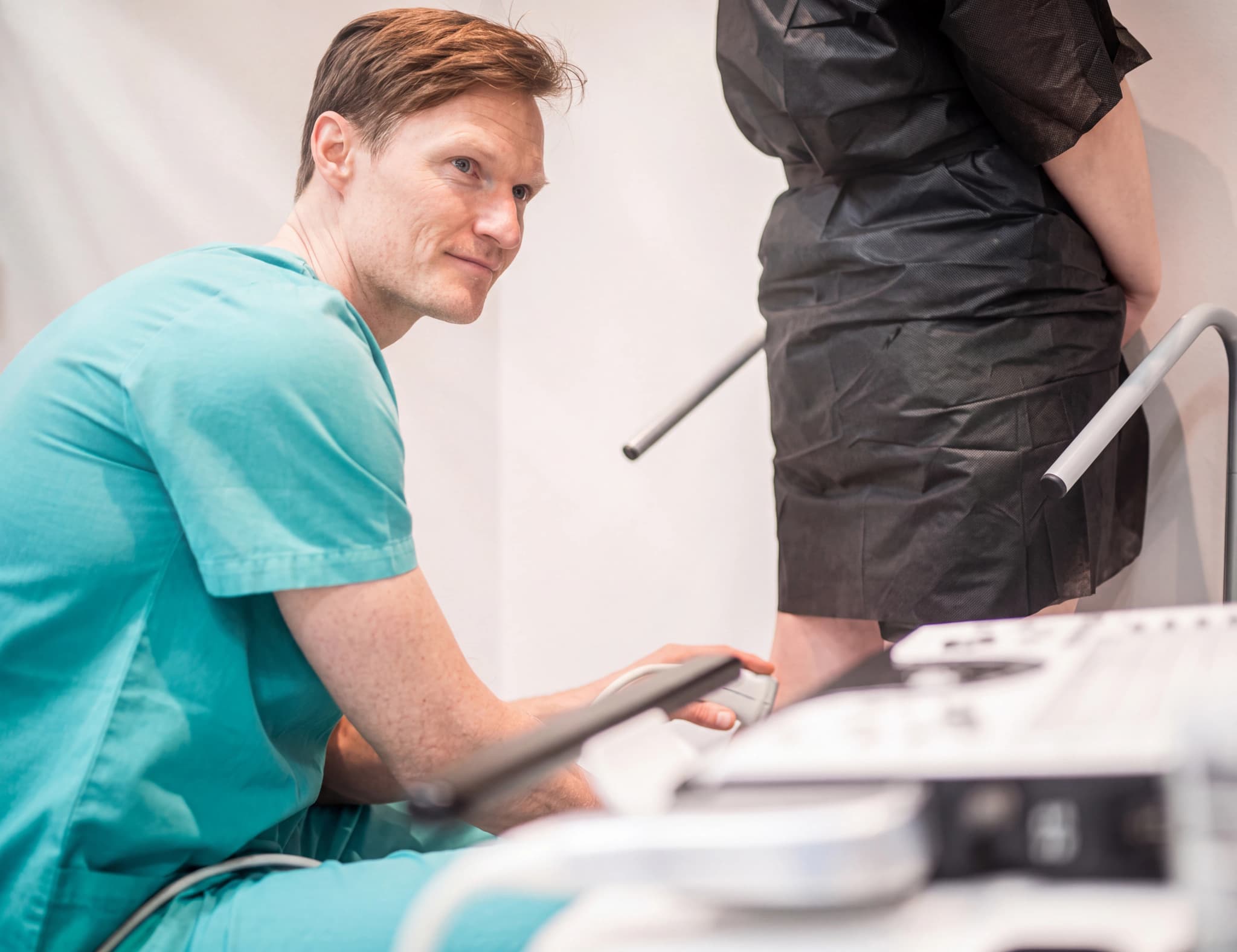 Läkare i ljusgröna arbetskläder utför en ultraljudsundersökning på en patients ben, medan patienten står med stöd.