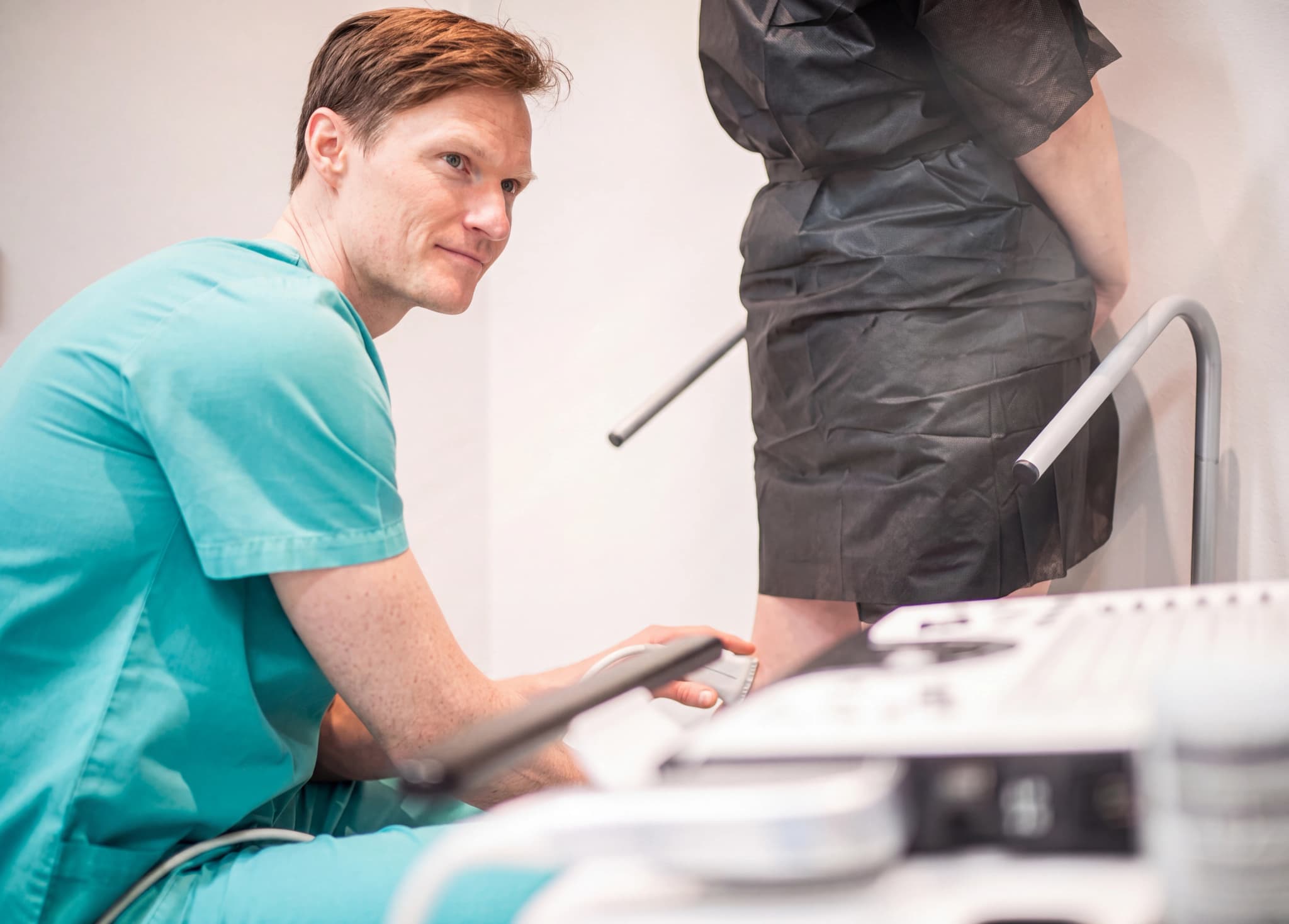 Läkare i grön arbetsklädsel utför en ultraljudsundersökning på en patients ben, med en ultraljudsapparat synlig i förgrunden.