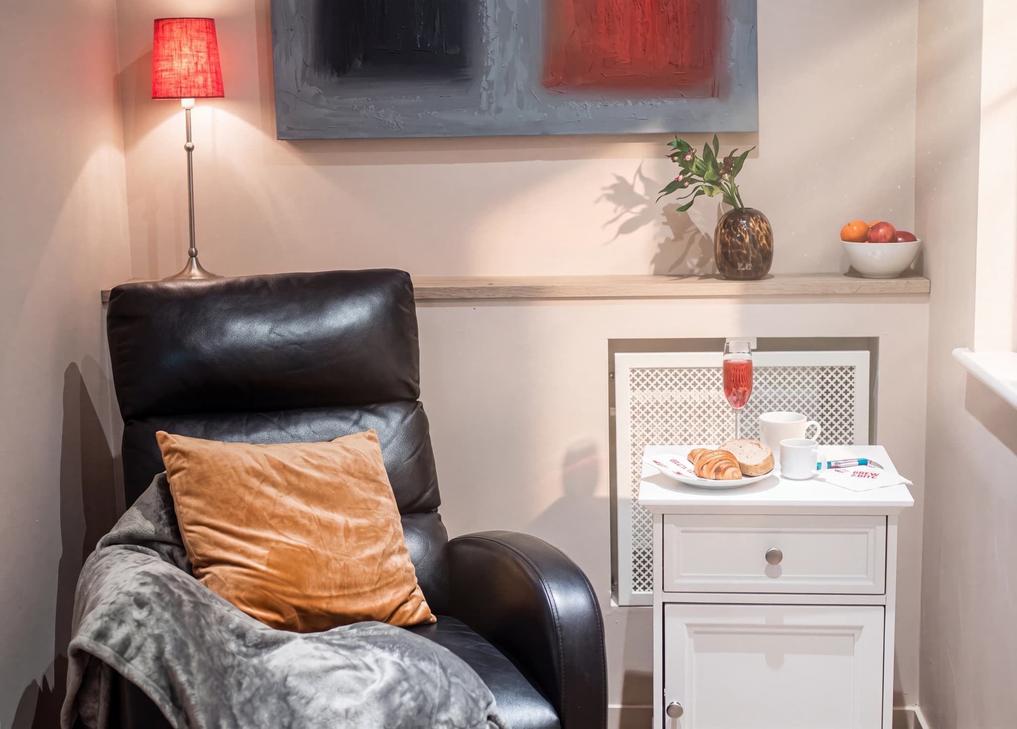 En svart läderfåtölj med en orange kudde och en grå filt, bredvid ett vitt sidobord med en frukostbricka, ett glas röd dryck och en vas med växter i bakgrunden.