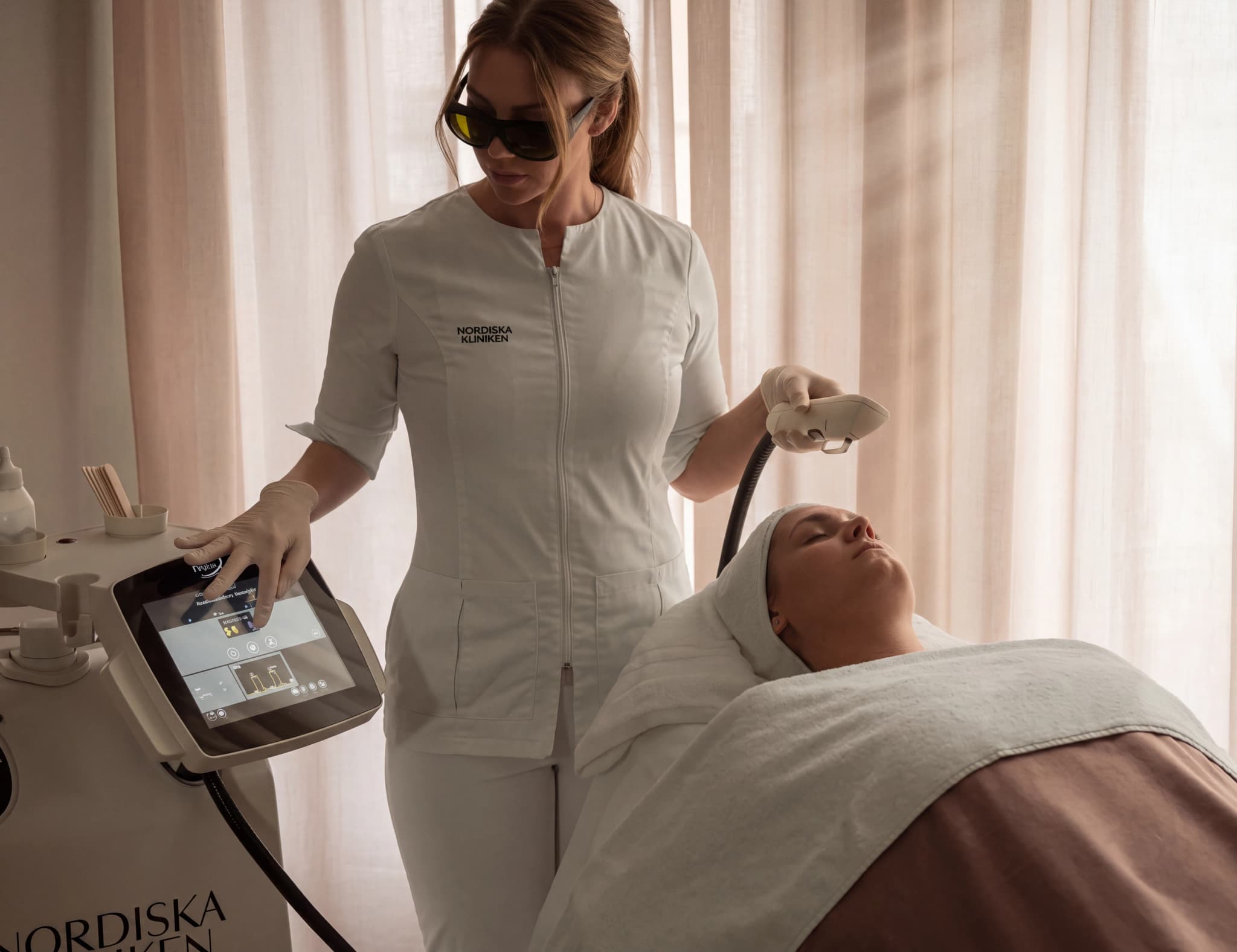 Estetiker i vit uniform med skyddsglasögon använder en lasermaskin på en patient som ligger på en brits.