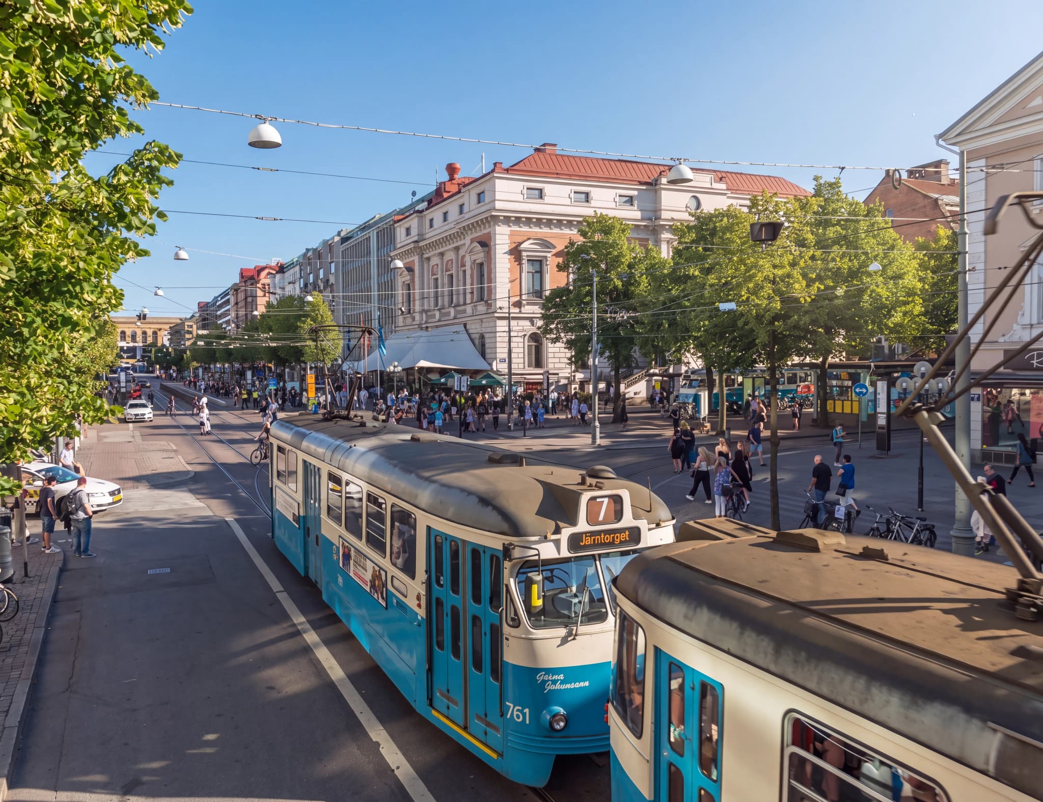 Blå och vita spårvagnar kör längs en livlig stadsgata med träd och byggnader i bakgrunden, många fotgängare på trottoaren.