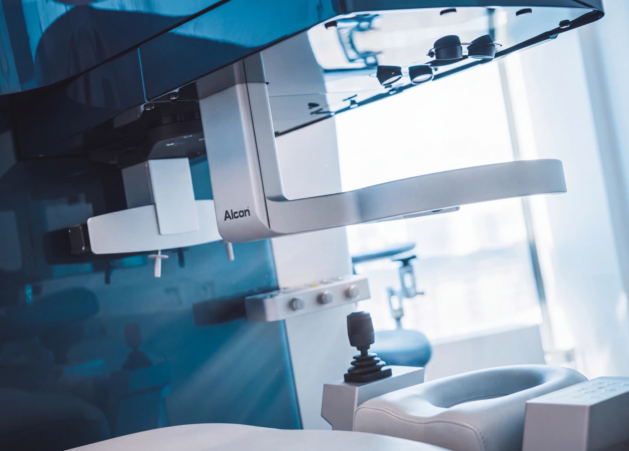 Närbild av en Alcon oftalmisk kirurgisk mikroskop utrustning i en ljus klinikmiljö, med fokus på instrument och en del av en behandlingsstol.