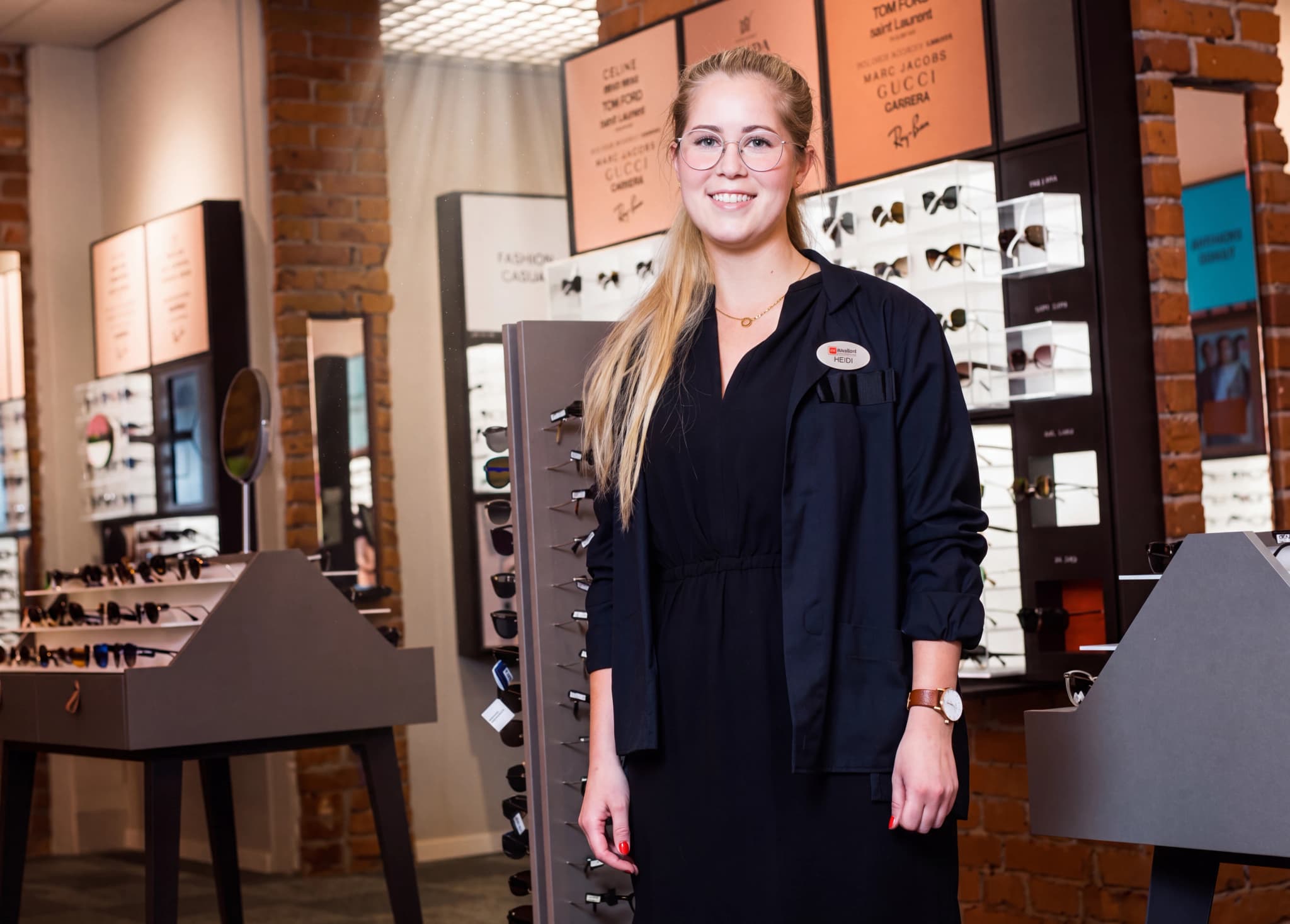 En leende kvinna med glasögon och arbetskläder assisterar i en optikerbutik.
