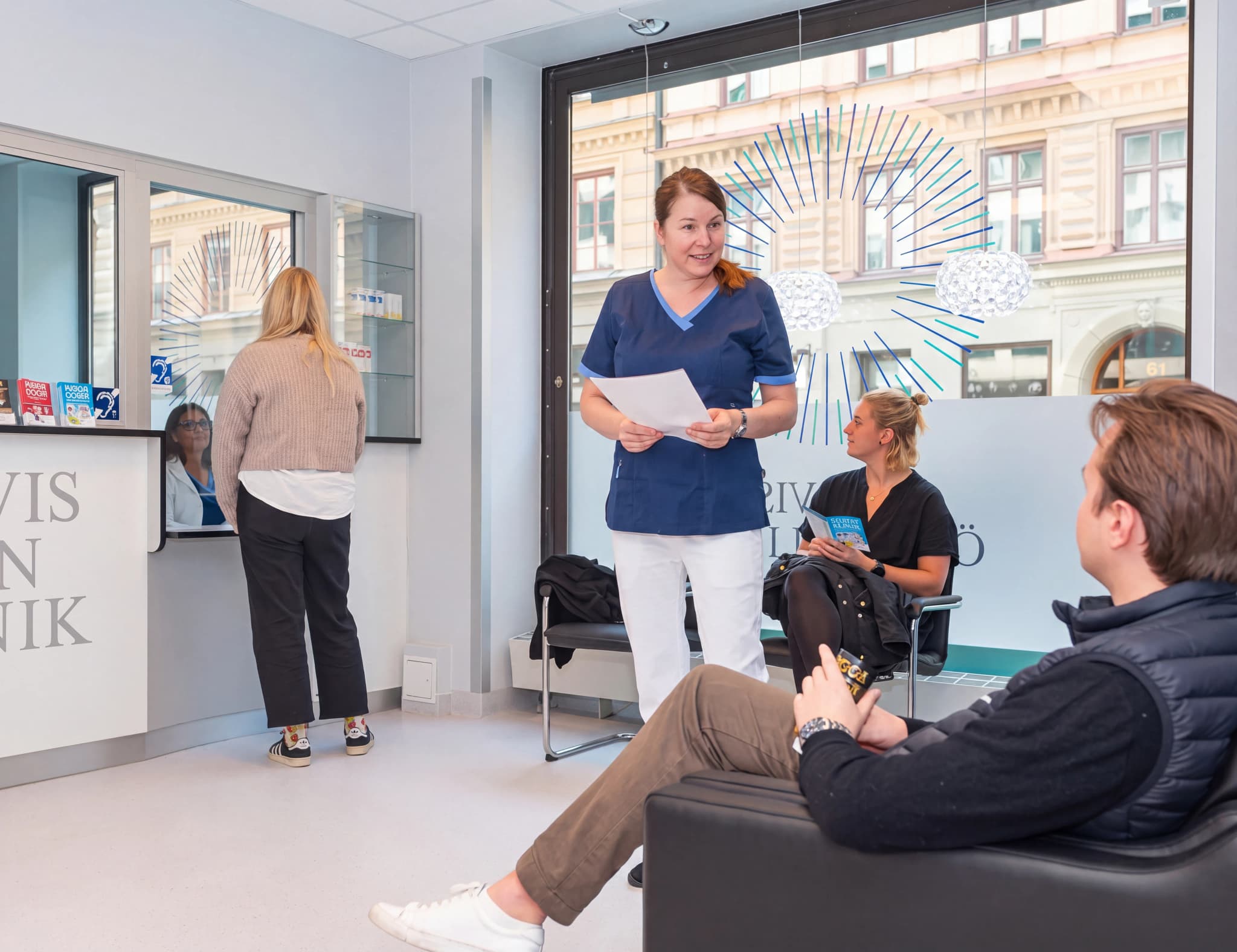 Receptionschef som hjälper en patient vid receptionen på en klinik, med en annan patient sittande i väntrummet.