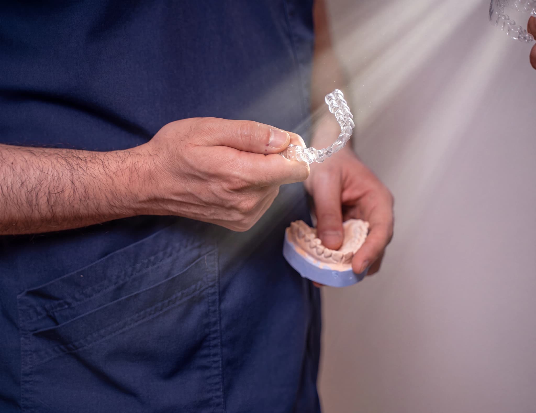 Tandläkare som håller en genomskinlig tandställning (aligner) och en gipsavgjutning av tänder i en hand, med en annan person i bakgrunden som håller en liknande aligner.