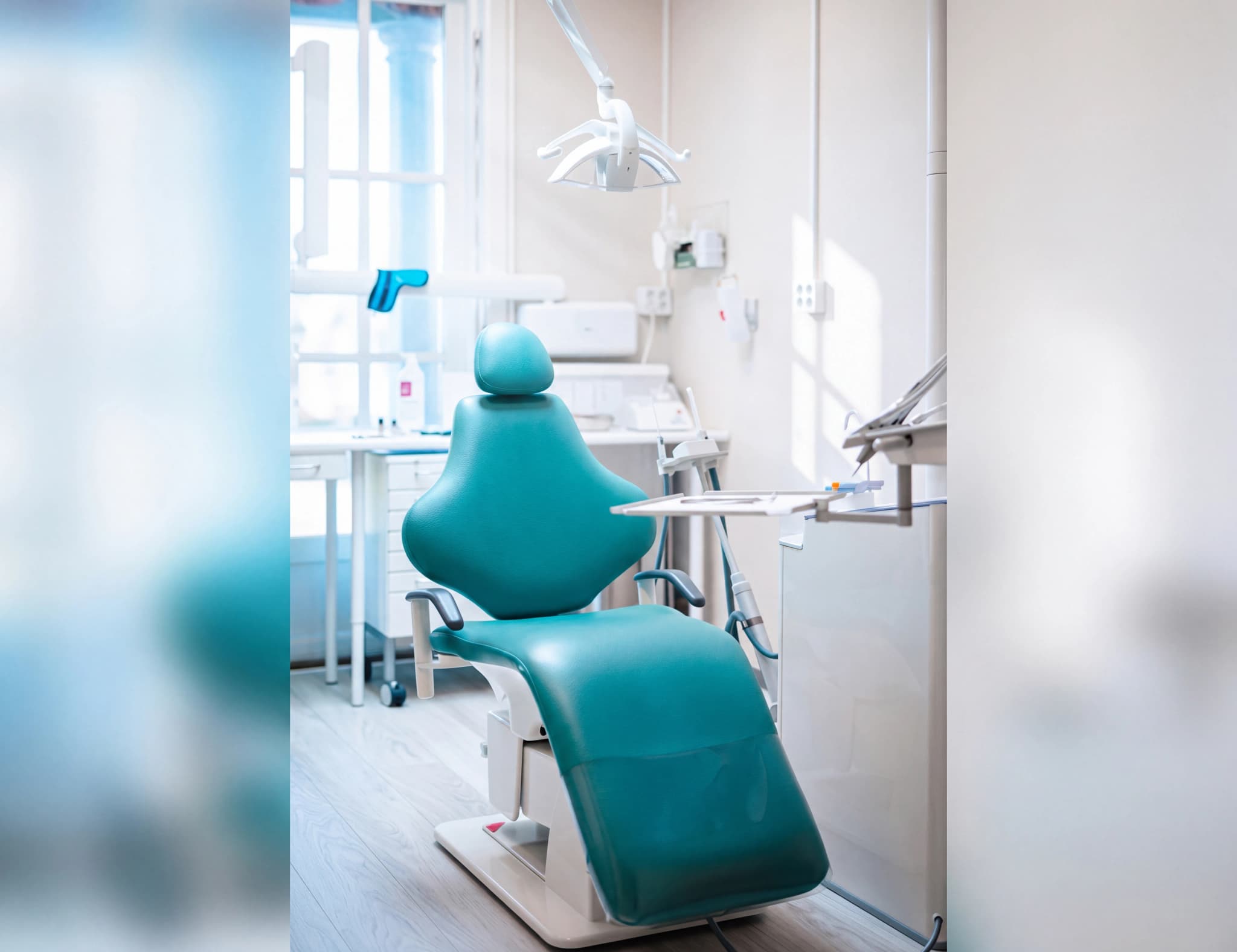 En grönblå dentalstol står i ett ljust undersökningsrum med ett fönster i bakgrunden.