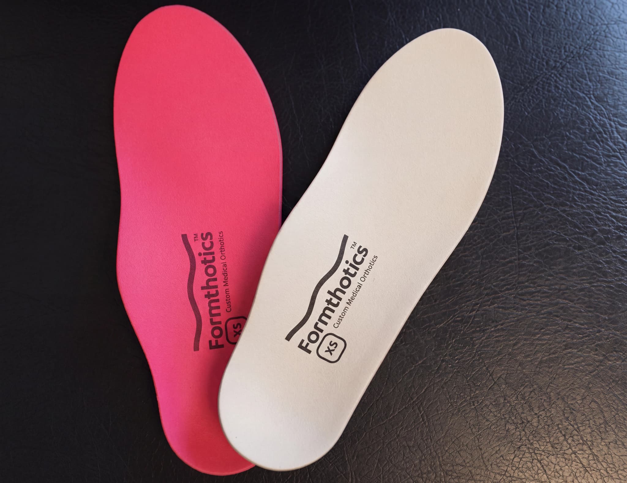 Ett par ortopediska iläggssulor, en i starkt rosa och en i vit, liggande på en mörk yta. Båda har texten "Formthotics Custom Medical Orthotics" tryckt på sig.