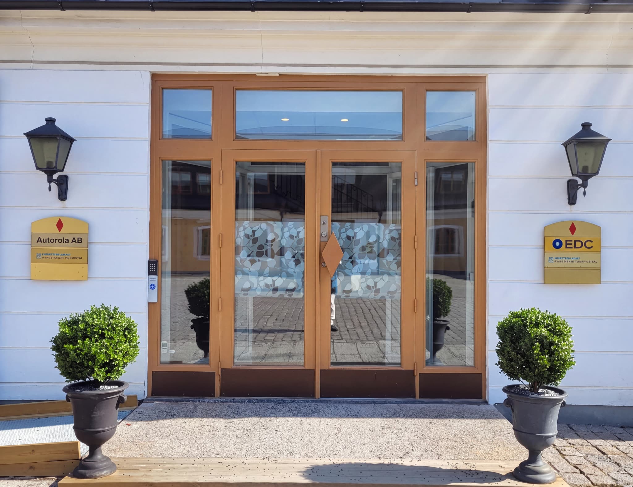Byggnadsingång med dubbeldörrar, fönster, en skylt med texten "Autorola AB" till vänster och en skylt med logotypen "EDC" till höger. Två krukväxter står på var sin sida om entrén.