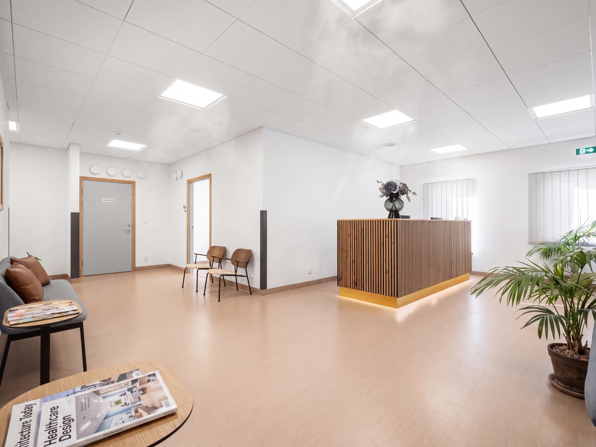 En mottagningsdisk med träribbor och en gyllene bas i en ljus och modern klinik, med väntrumsstolar och en dörr i bakgrunden.