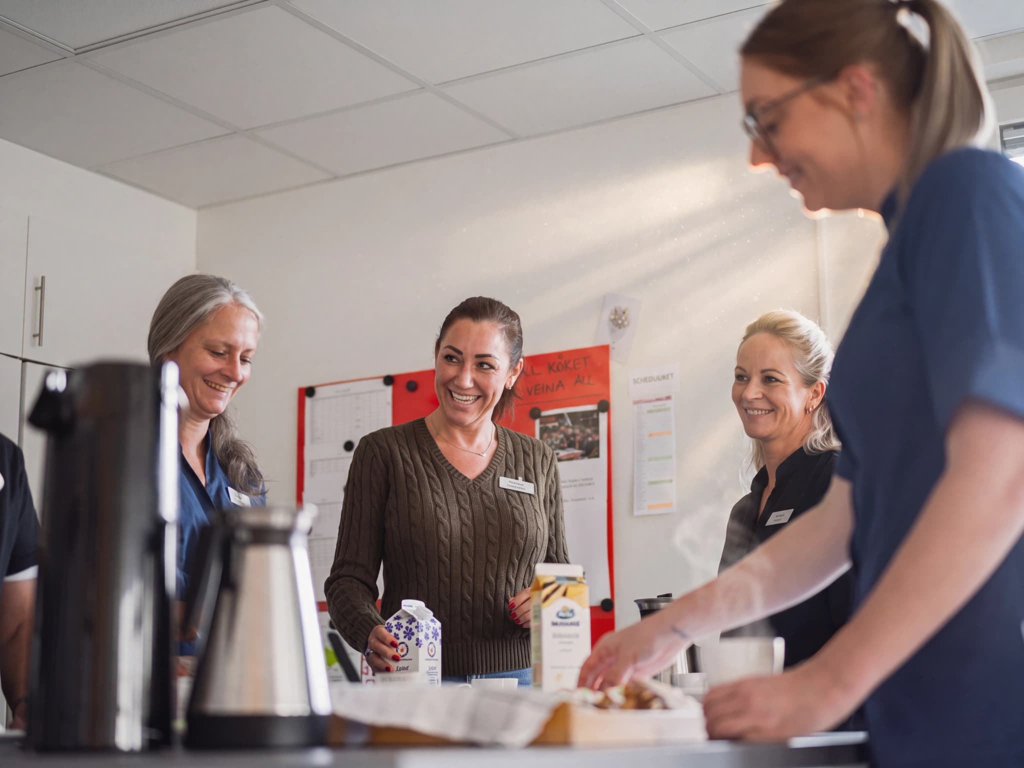 Fyra kvinnliga kollegor ler och pratar runt ett kaffebord i ett ljust kontorsutrymme.