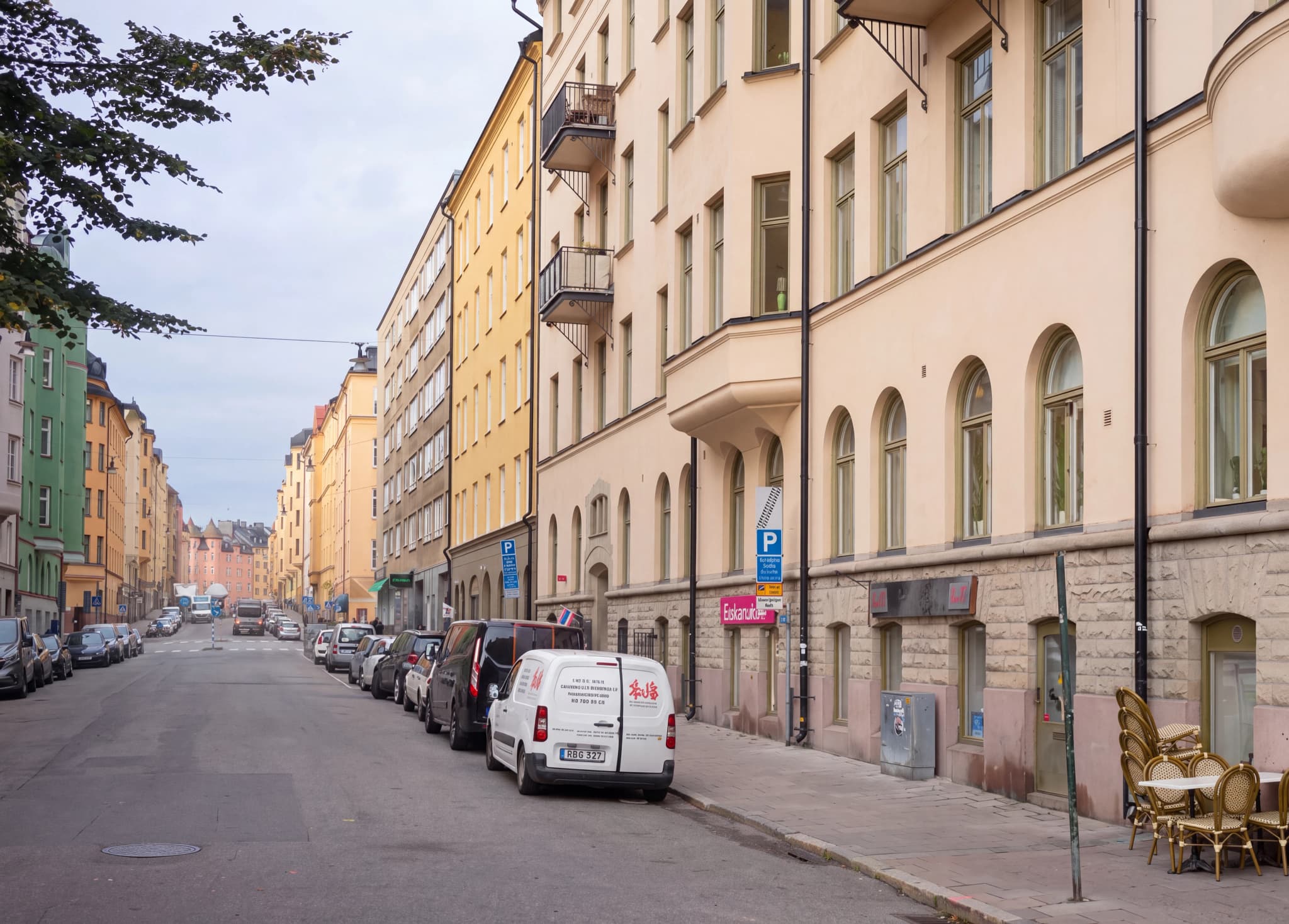 Vy över en gata med flerfamiljshus i olika färger och parkerade bilar längs trottoaren.