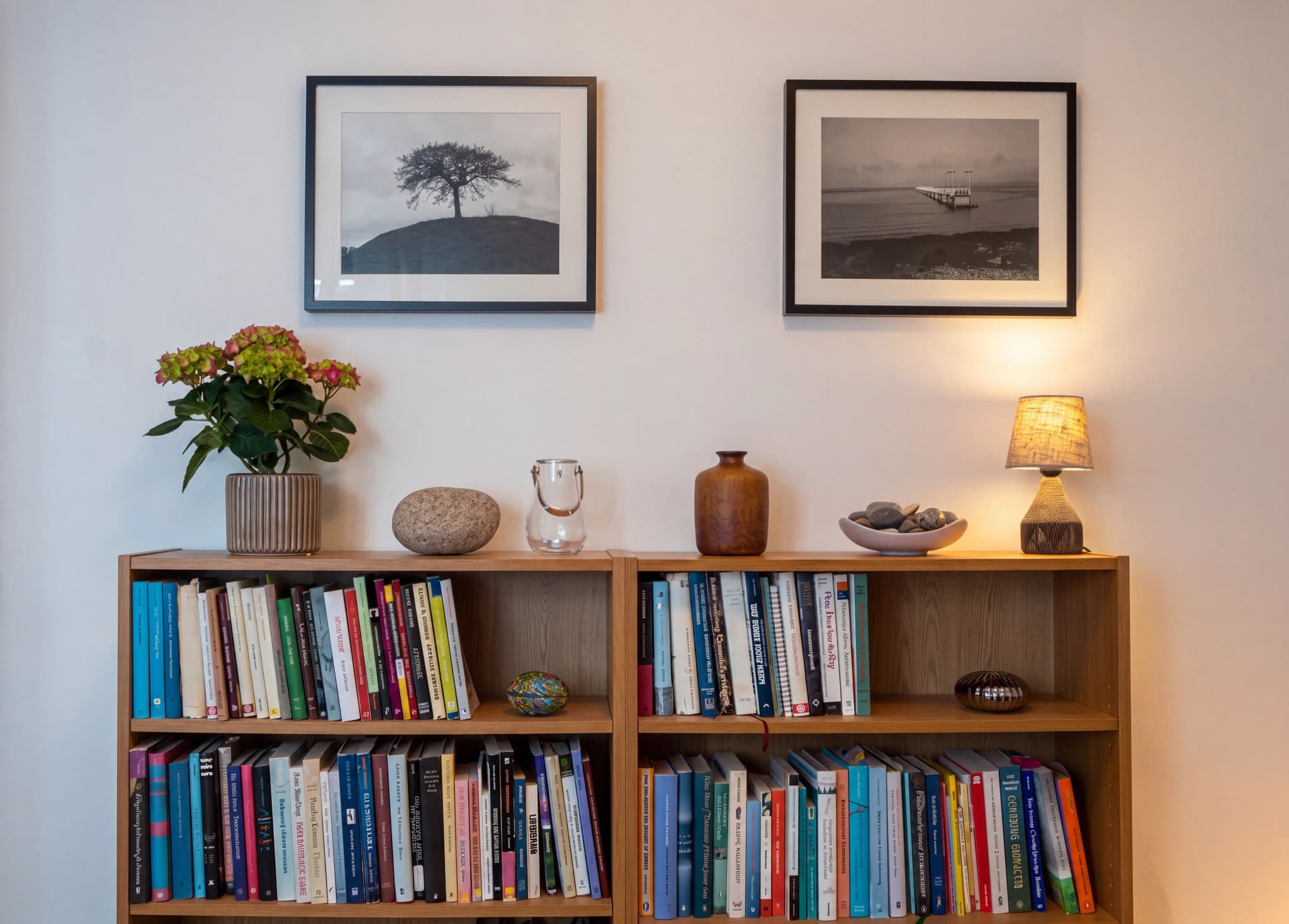 En bokhylla i trä fylld med böcker, prydnadssaker som en vas och stenar, samt en liten bordslampa på en av hyllorna. Ovanför bokhyllan hänger två svartvita fotografier på väggen.