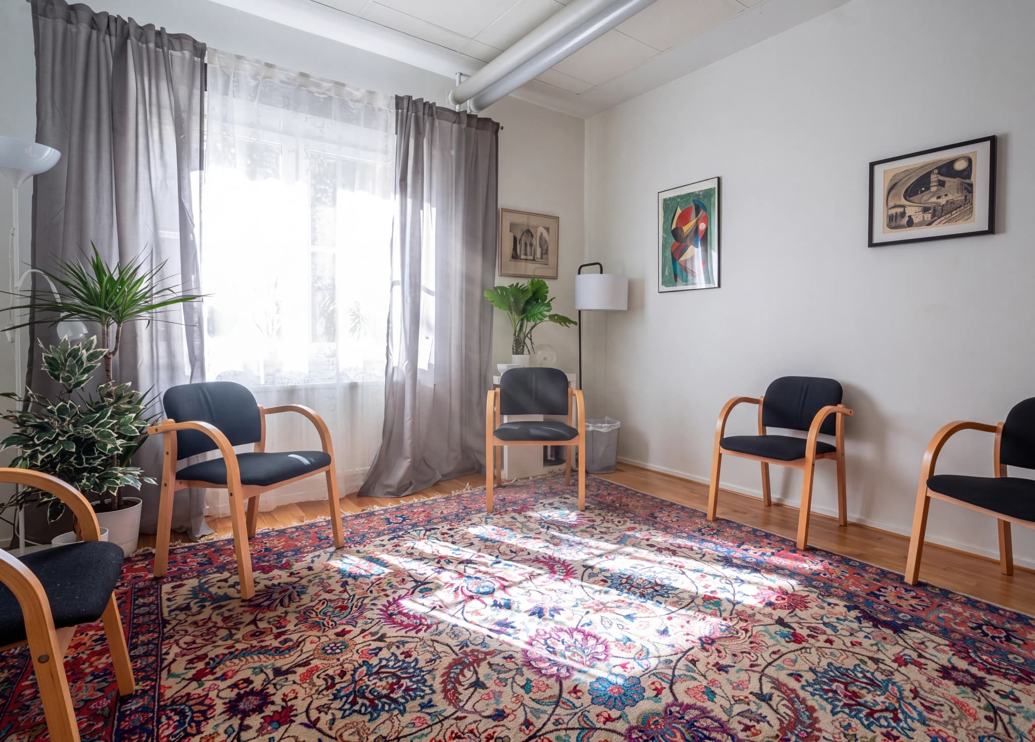 Ett väntrum med svarta stolar med trästomme, en stor orientaliskt mönstrad matta och fönster med grå gardiner.