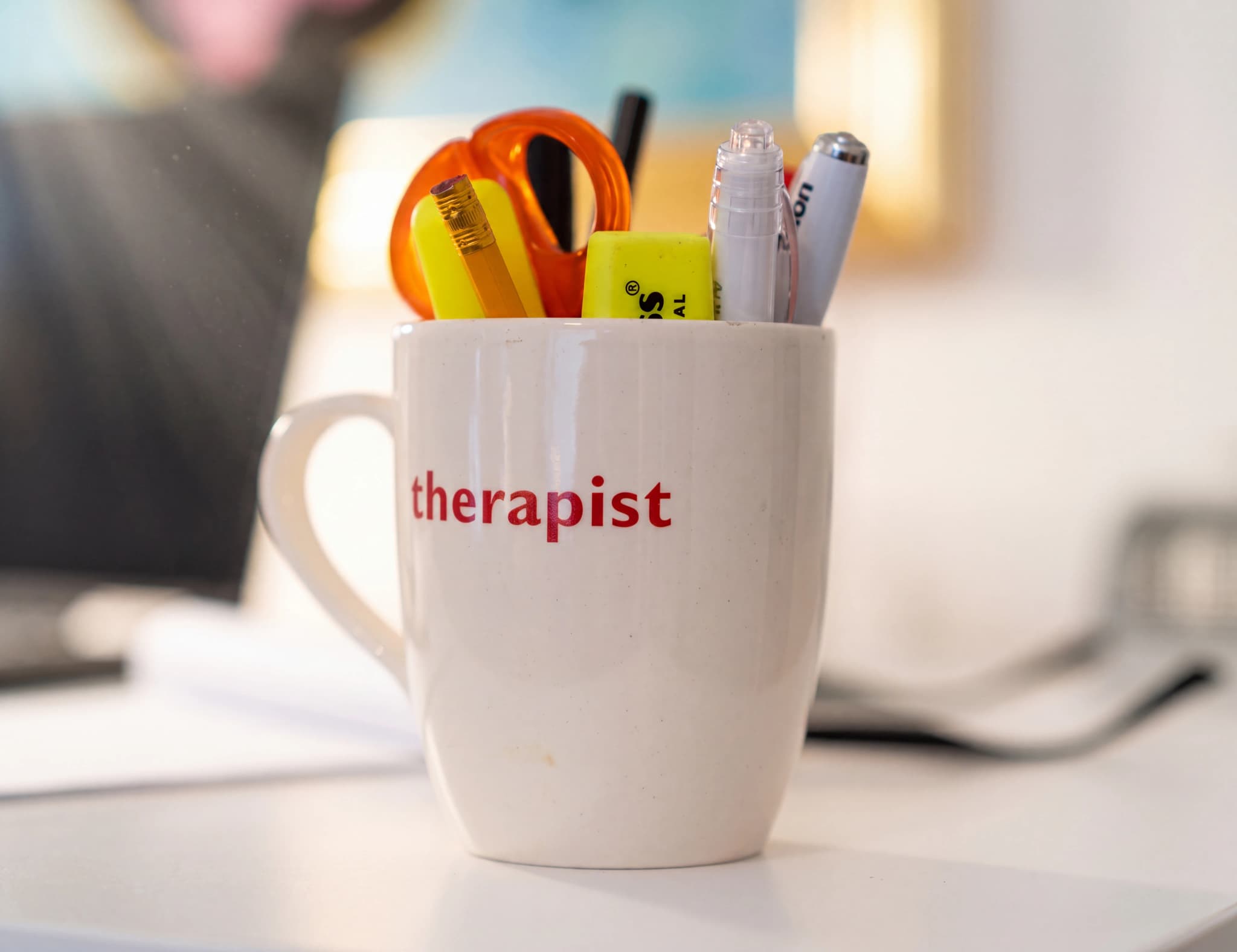 En vit mugg med texten "therapist" i rött tryck, fylld med sax, pennor och en överstrykningspenna, står på ett vitt skrivbord.