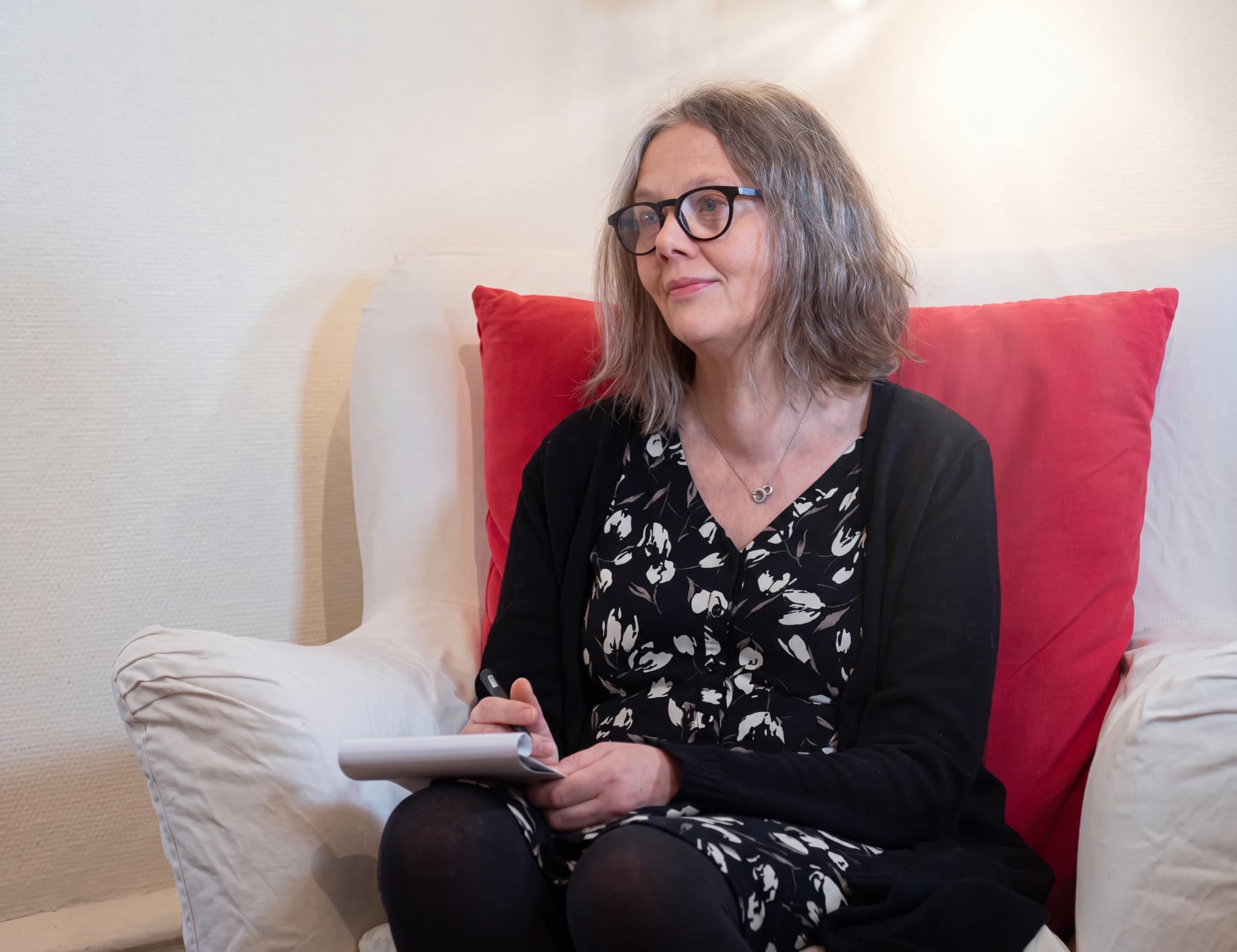 Kvinna med glasögon sitter i en stol med en anteckningsbok i händerna.