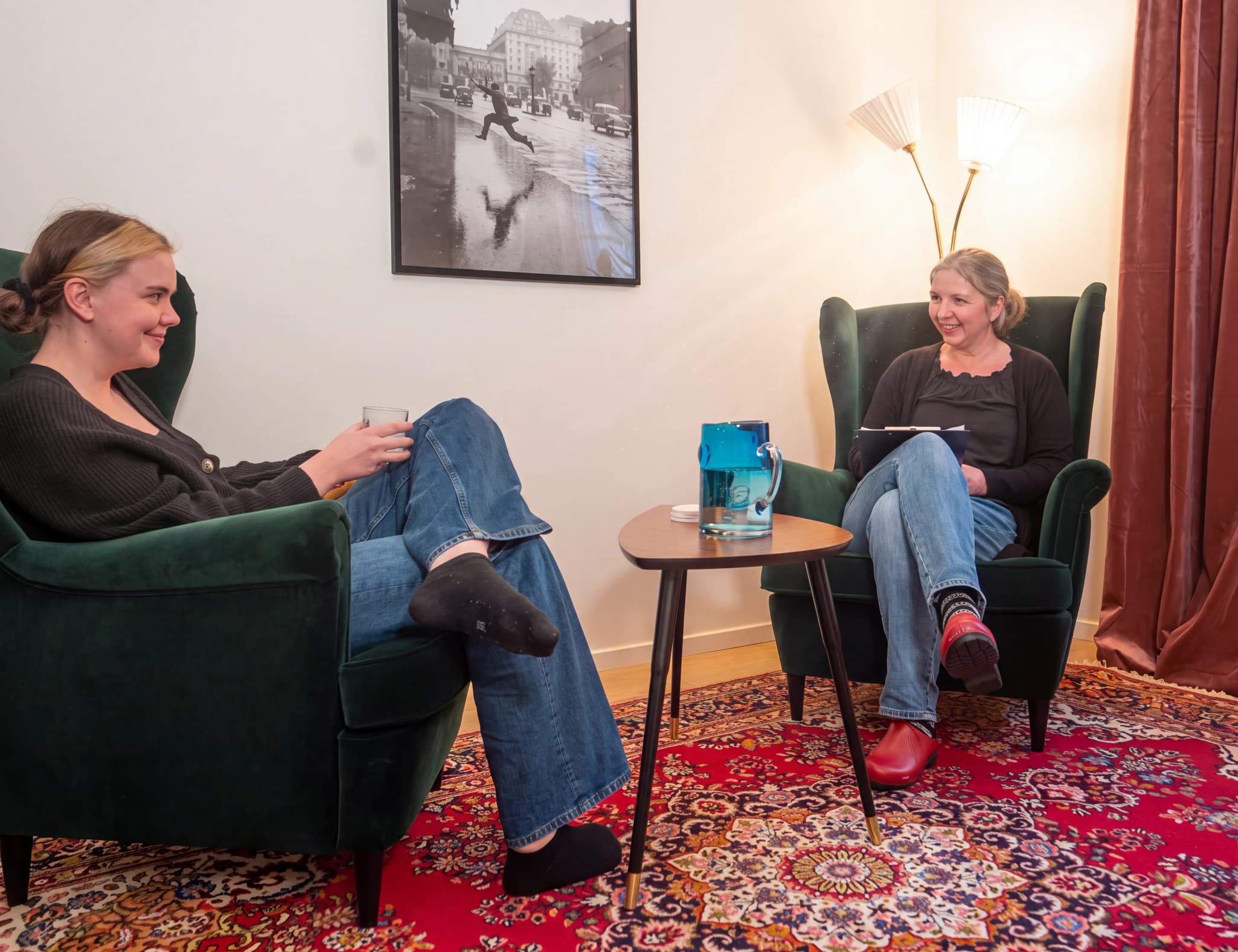 En terapeut pratar med en patient som sitter i en fåtölj i ett ljust och välkomnande terapirum med en matta och konst på väggen.