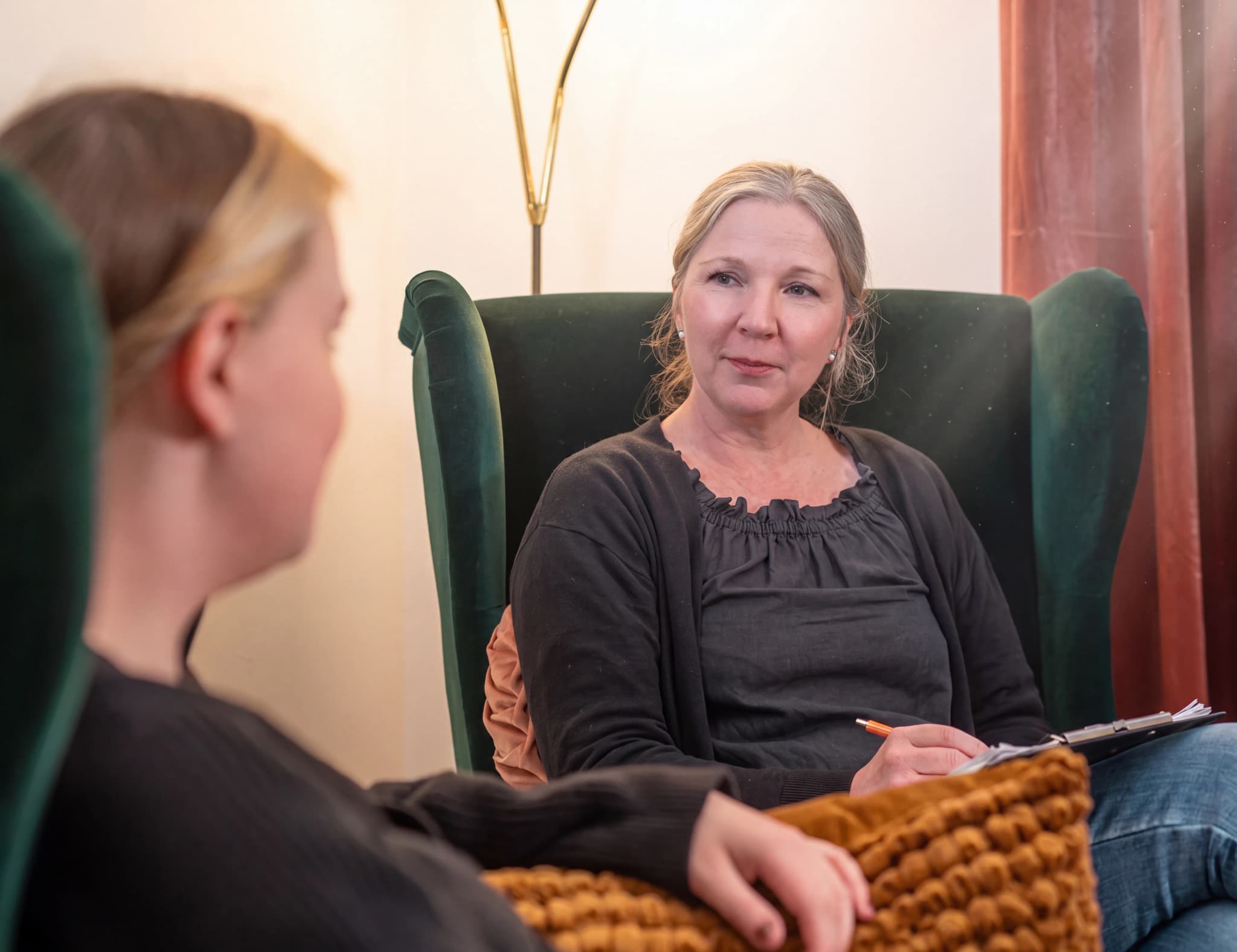 En terapeut sitter i en mörkgrön fåtölj och för anteckningar på ett block, medan hon tittar på en patient som sitter i en annan fåtölj och är vänd bort från kameran.