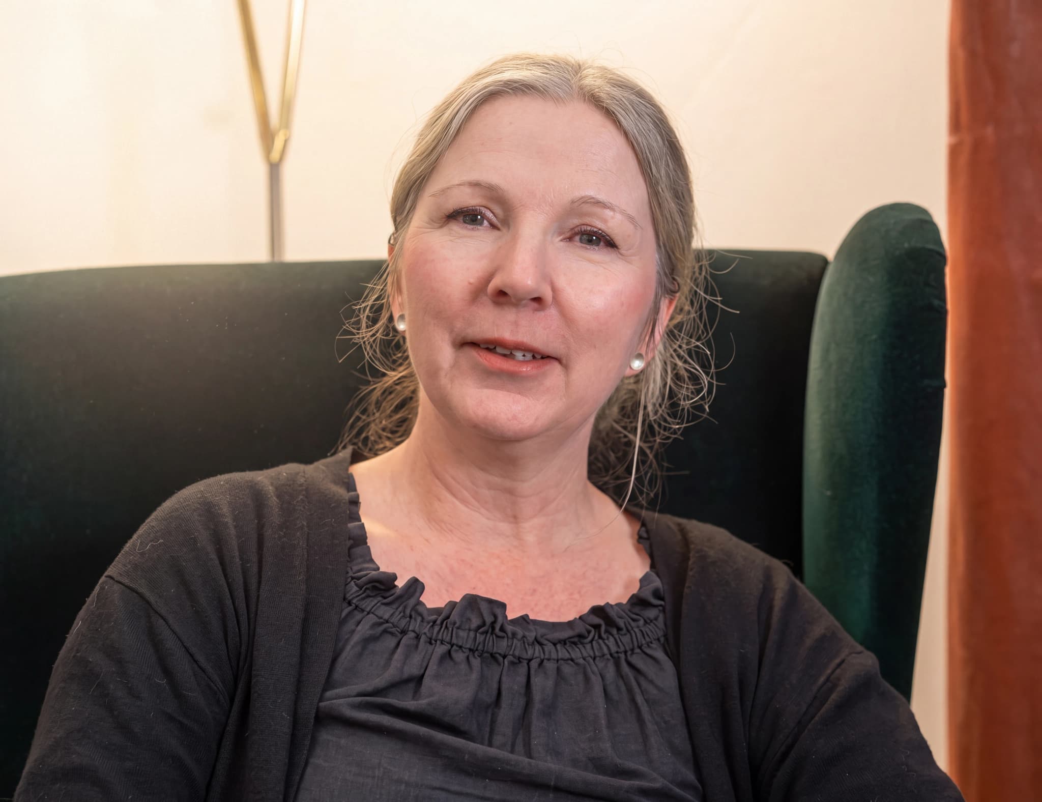 Kvinna med grånande hår sitter i en mörkgrön fåtölj och ler.