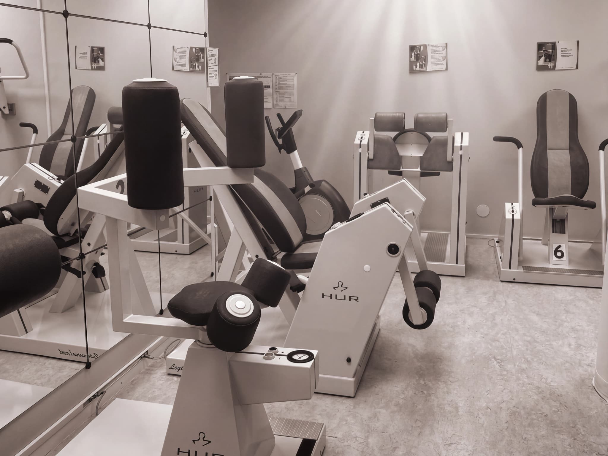 Färgbild i sepia av ett rehabiliteringsgym med flera träningsmaskiner av märket HUR, inklusive sittande maskiner och liggande maskiner.