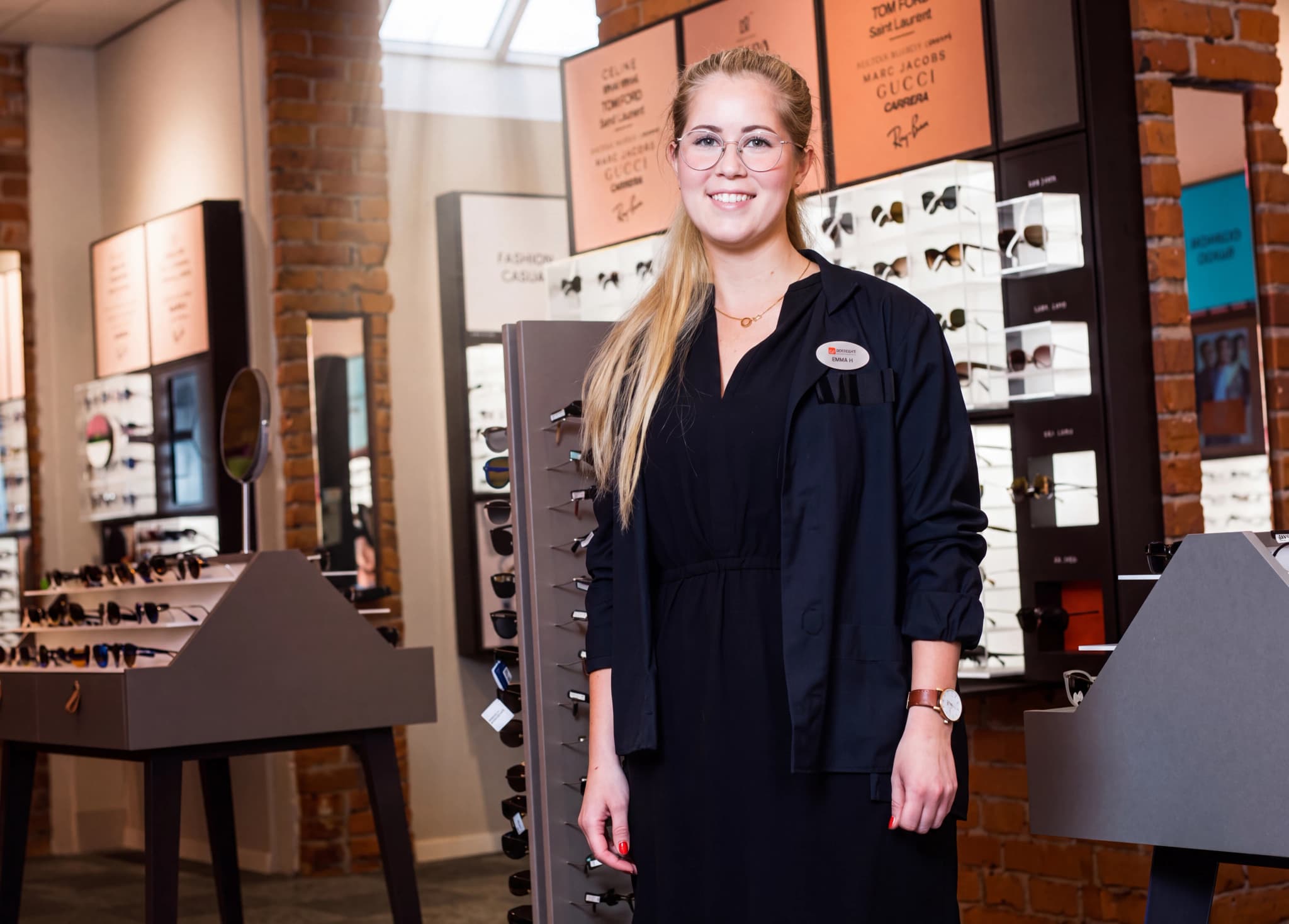 En ung kvinna med glasögon och blont hår ler mot kameran i en butik som säljer glasögon, med montrar fyllda med glasögon i bakgrunden.
