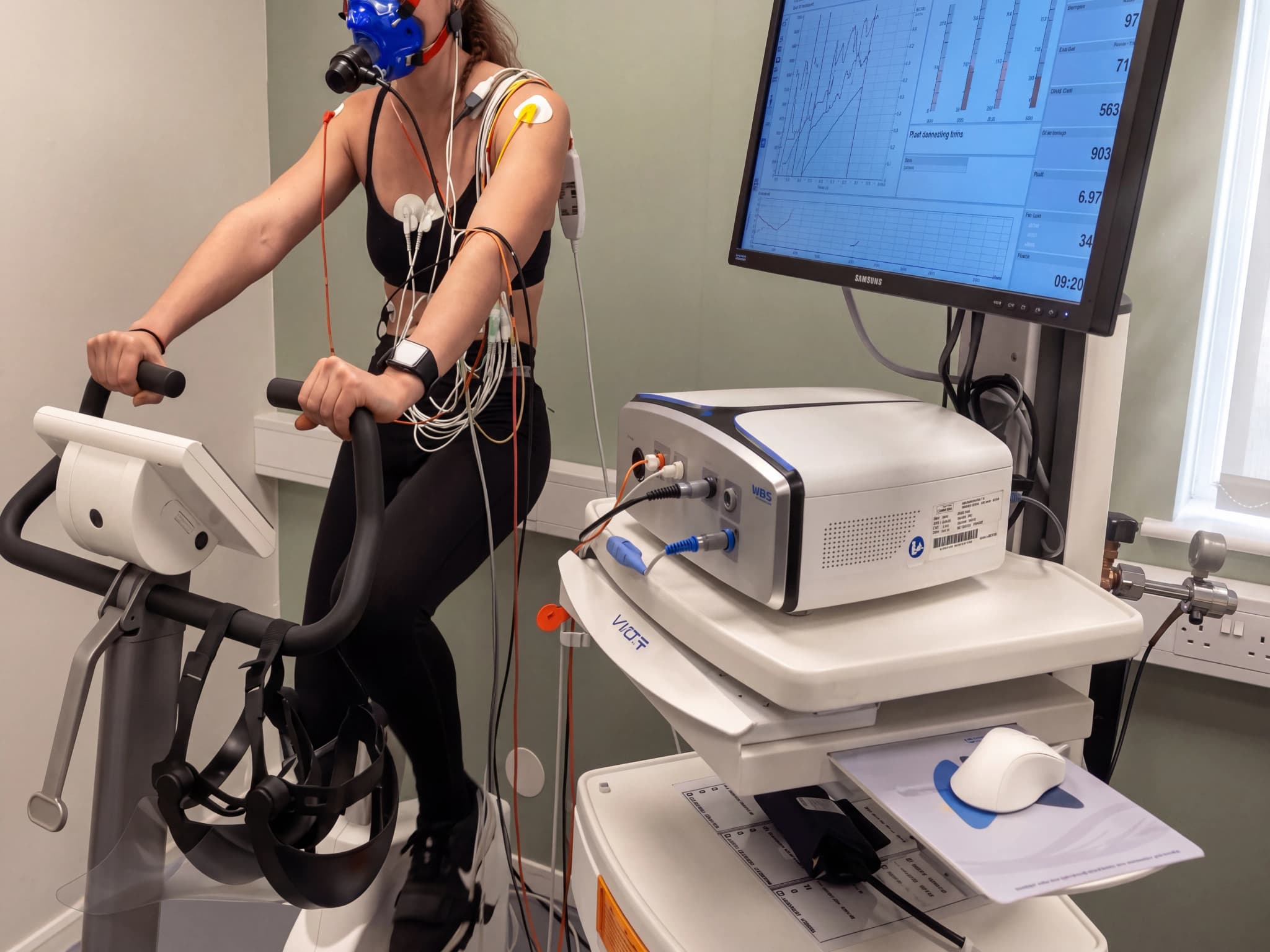 Kvinna utför ett konditionstest på en motionscykel med utrustning för att mäta hjärtfunktion och syreupptag.