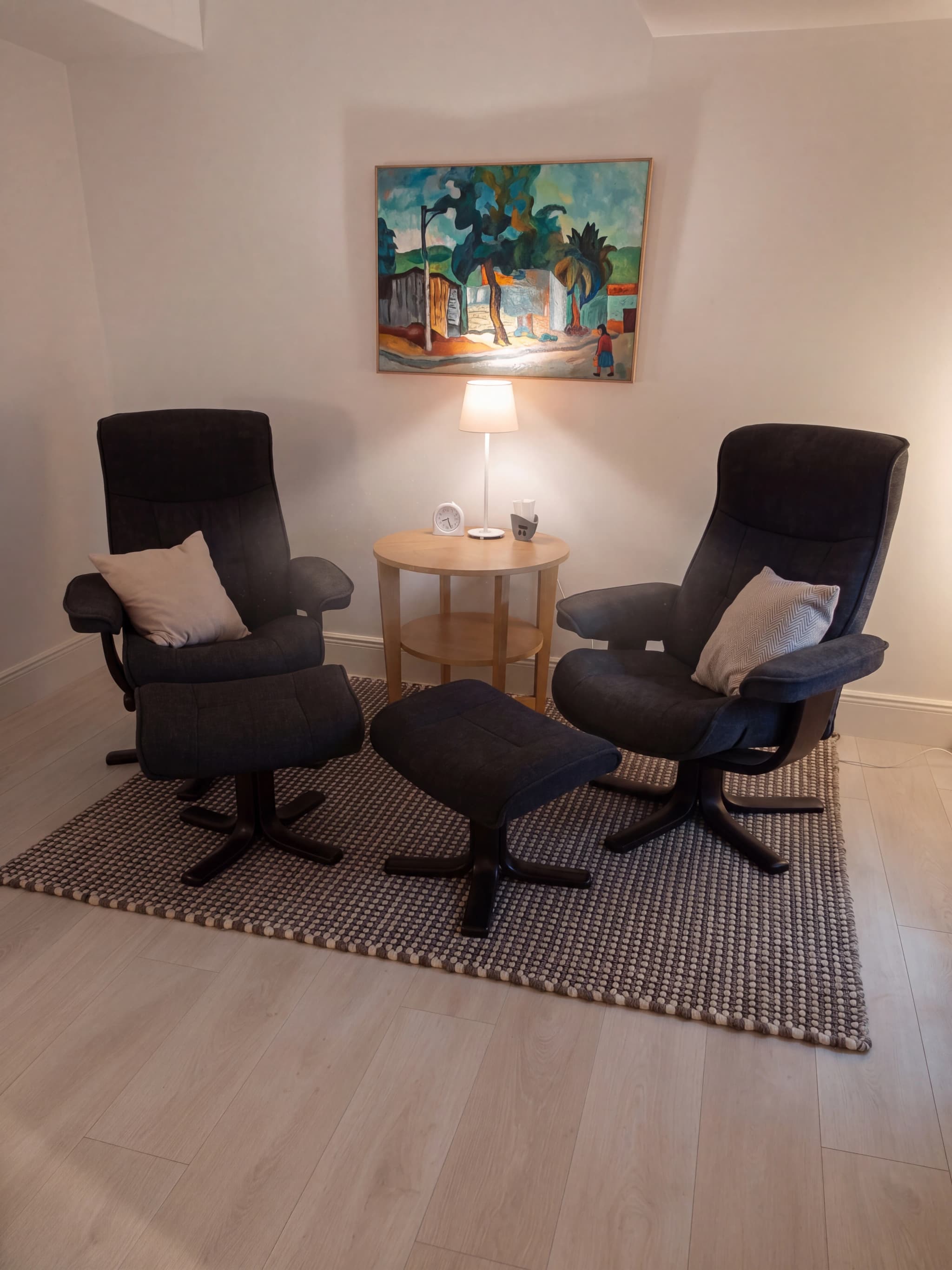 Två svarta reclinerfåtöljer med tillhörande fotpallar står på en matta i ett väntrum. En liten rund sidobord med en lampa och en väckarklocka står mellan dem. En färgstark tavla hänger på väggen ovanför.