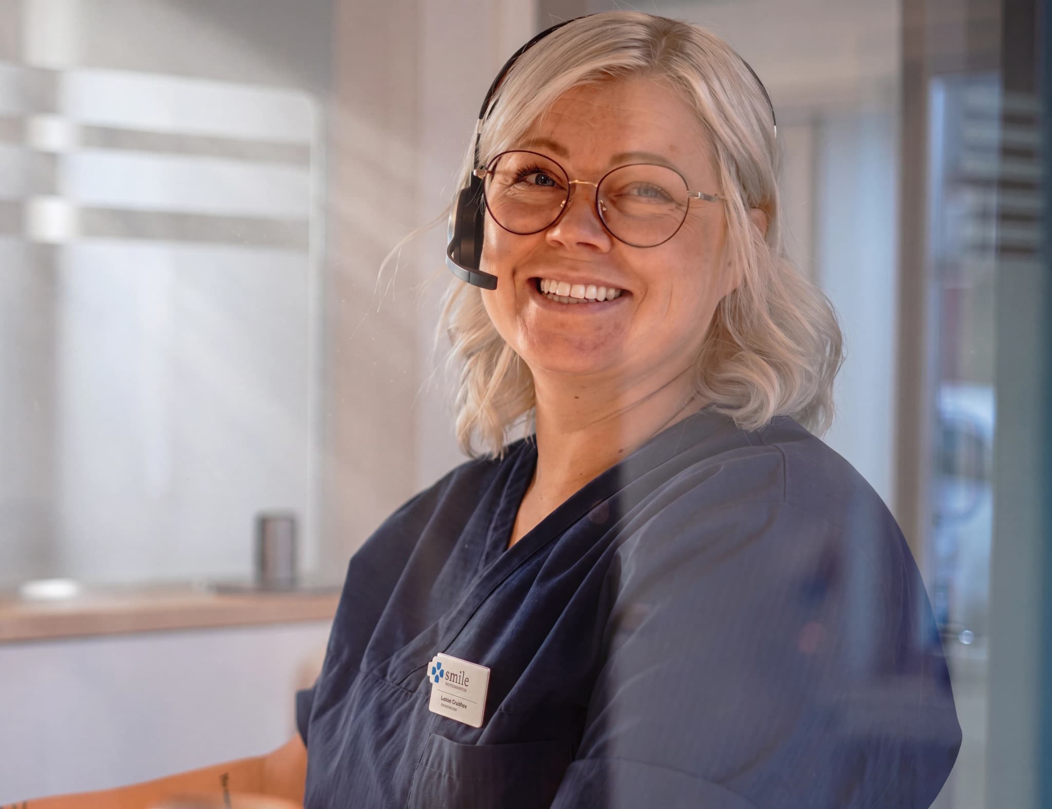 En kvinna med glasögon och headset ler mot kameran medan hon arbetar bakom en reception i en klinik.
