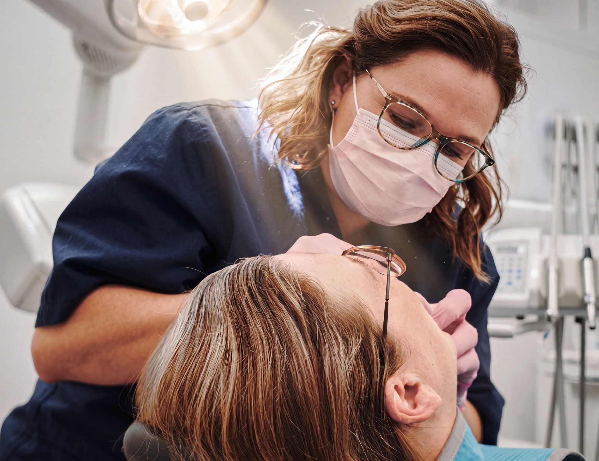Tandläkare med munskydd och glasögon undersöker en patients mun med ett instrument i en tandläkarmottagning.