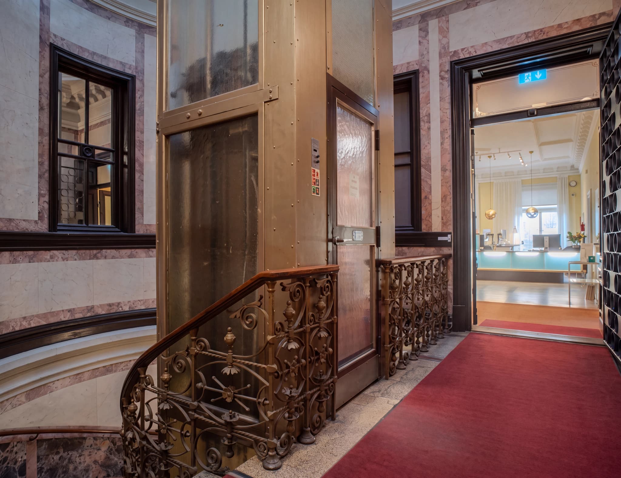Ett gammalt, utsmyckat hisschakt i metall med glaspaneler bredvid en svängd trappa med vackert smidesräcke. En röd heltäckningsmatta leder in i en ljus reception genom en öppen dörröppning.
