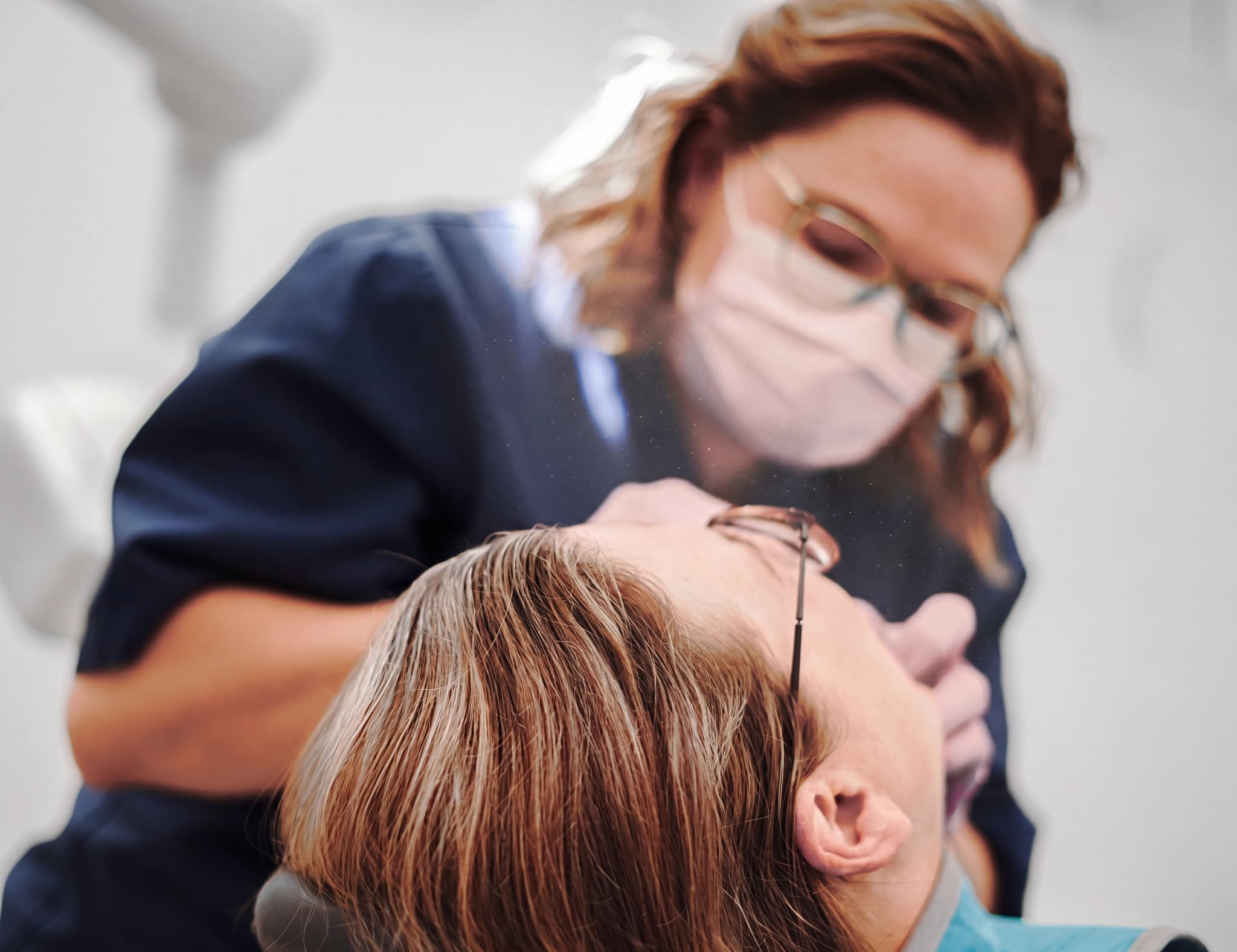 Tandläkare i mask och glasögon undersöker en patients mun med ett instrument.