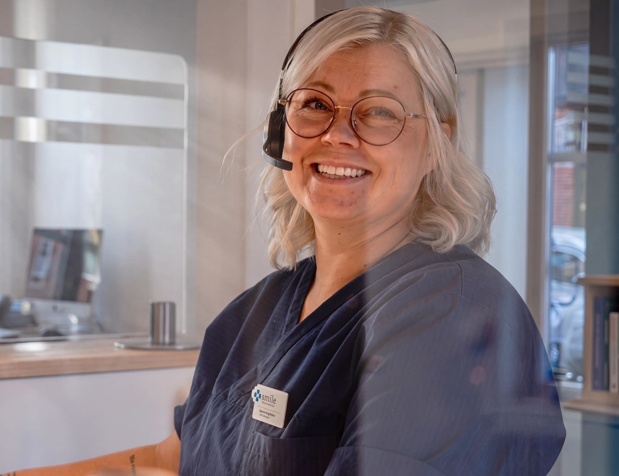 En kvinna med glasögon och headset ler in i kameran, klädd i mörkblå kläder med en namnskylt.