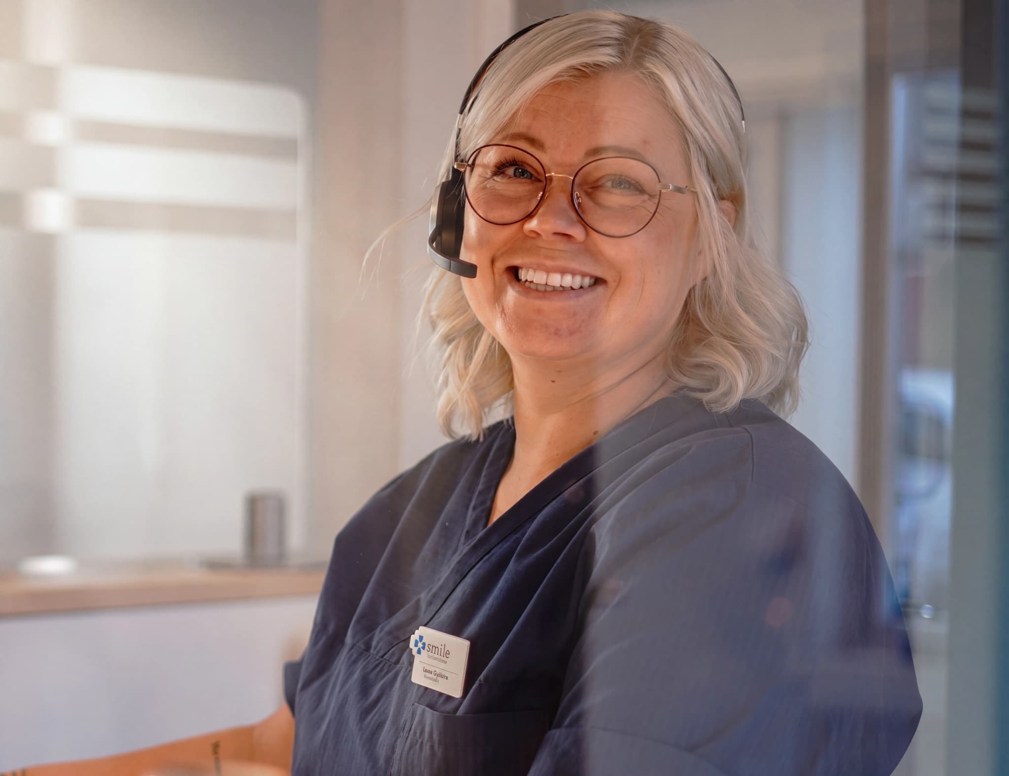 En kvinna med glasögon och headset ler medan hon arbetar vid receptionen på en klinik.