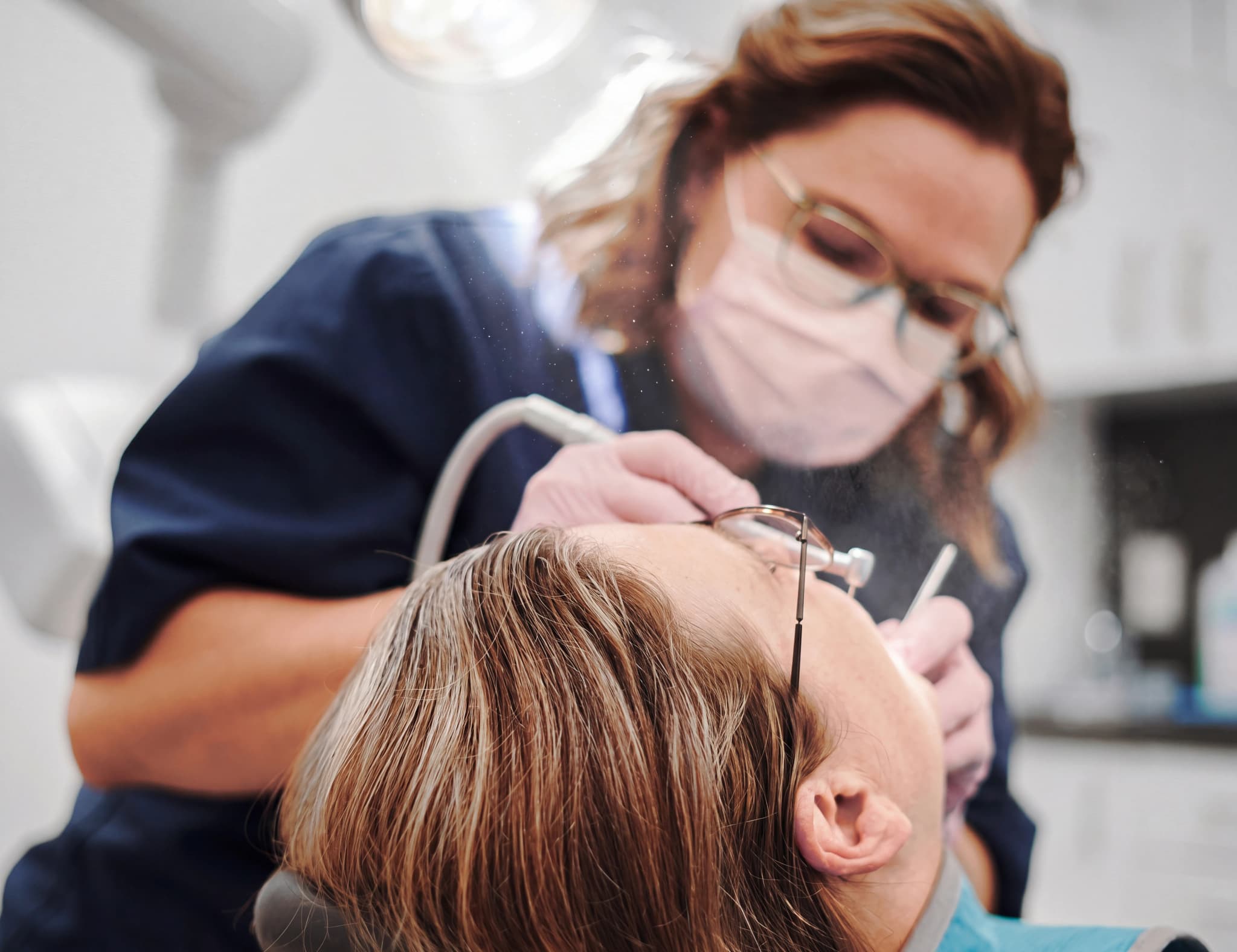 Tandläkare med munskydd och glasögon undersöker en patients mun med instrument.