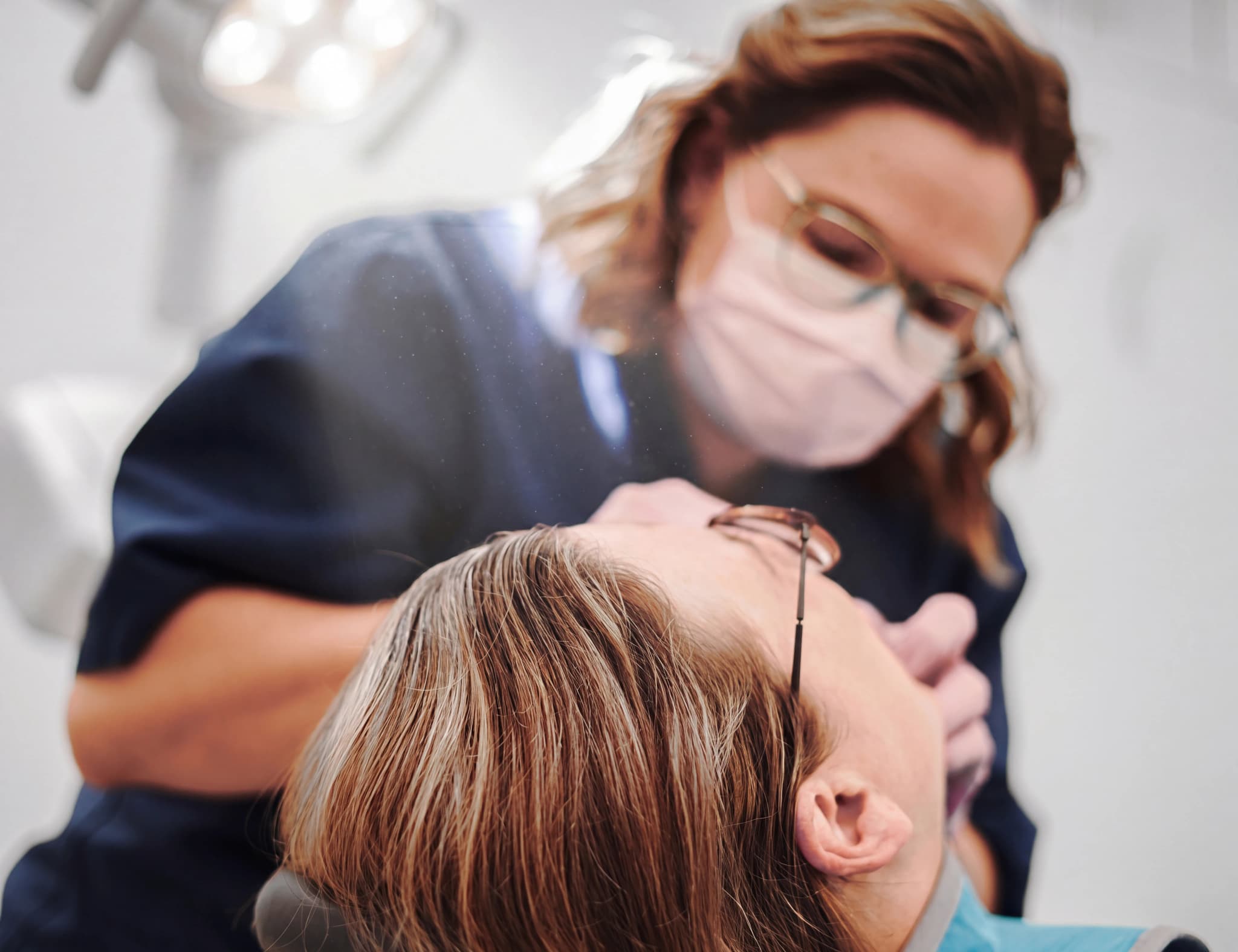 Tandläkare med munskydd och glasögon undersöker patientens mun med ett instrument.