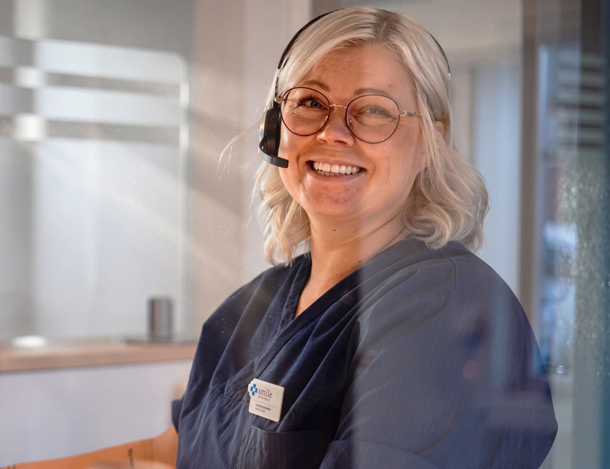 En kvinna med glasögon och headset ler mot kameran, klädd i mörkblå arbetskläder med en namnskylt.