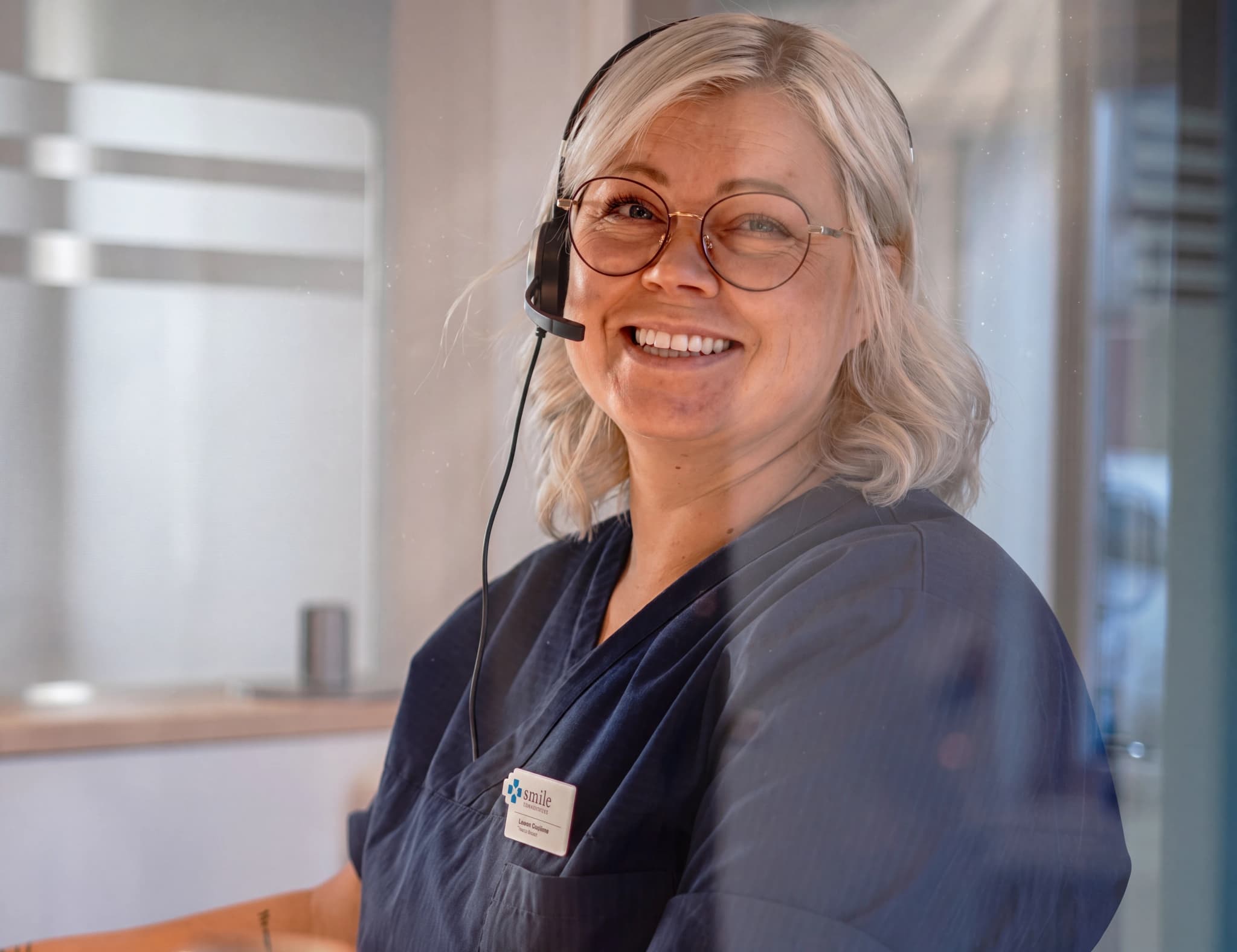 En kvinna med glasögon och headset ler mot kameran vid ett receptionsdisk.