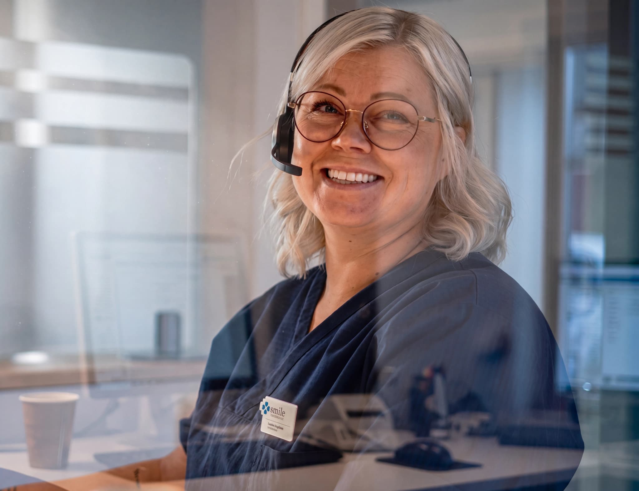 En leende kvinna med headset och glasögon som arbetar vid receptionen på en klinik.