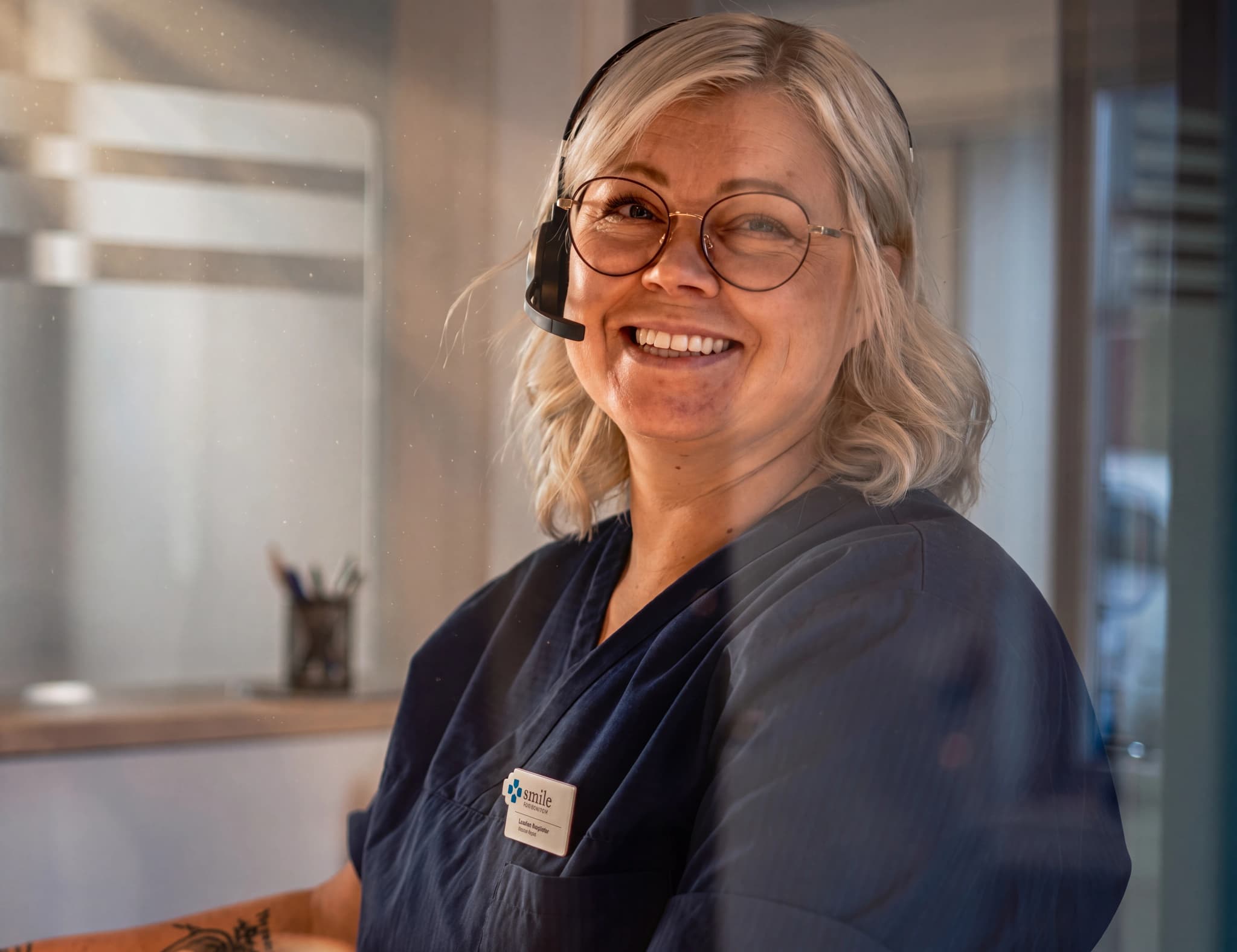 En kvinna med glasögon och headset ler mot kameran, iklädd mörkblå uniform med en namnskylt som visar texten "smile tandvård". Hon sitter troligen bakom en reception.