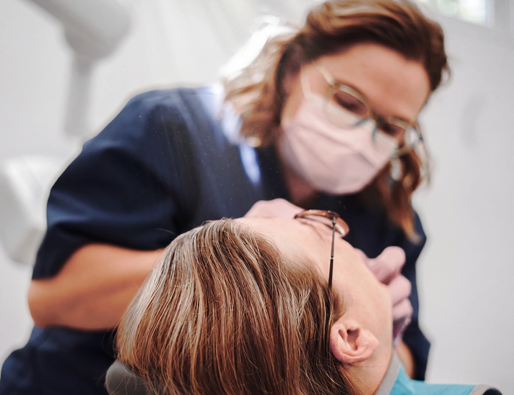 Tandläkare med munskydd och glasögon undersöker en patients mun med ett instrument i en klinik.