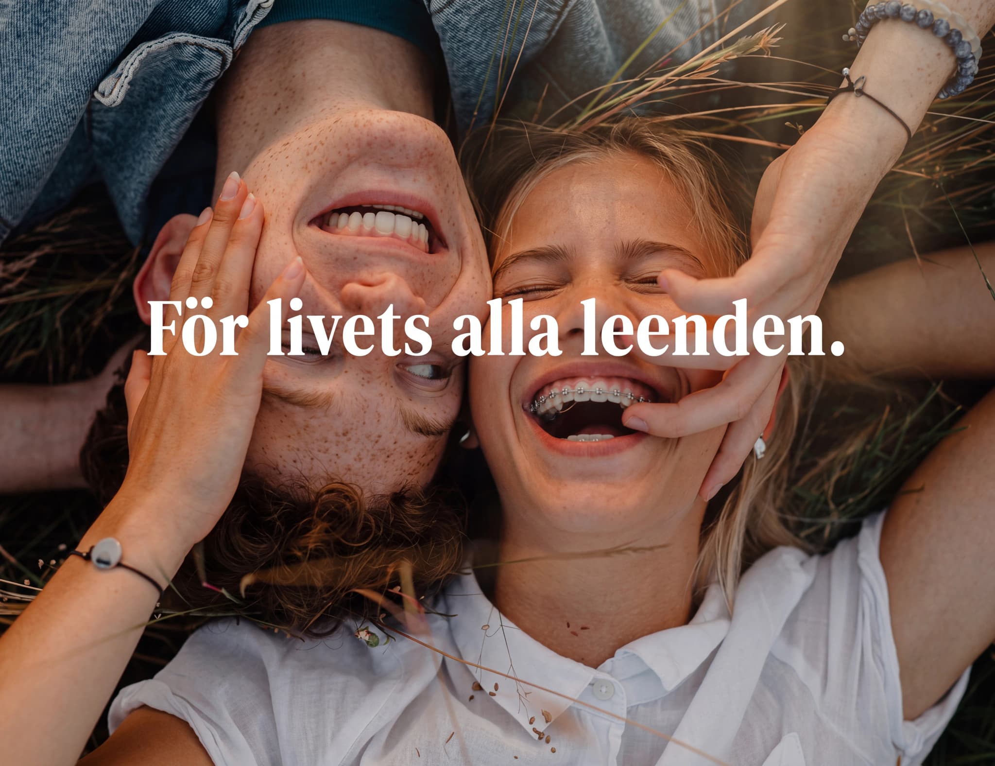 Ungt par med armarna runt varandra, skrattar lyckligt utomhus bland gräs.